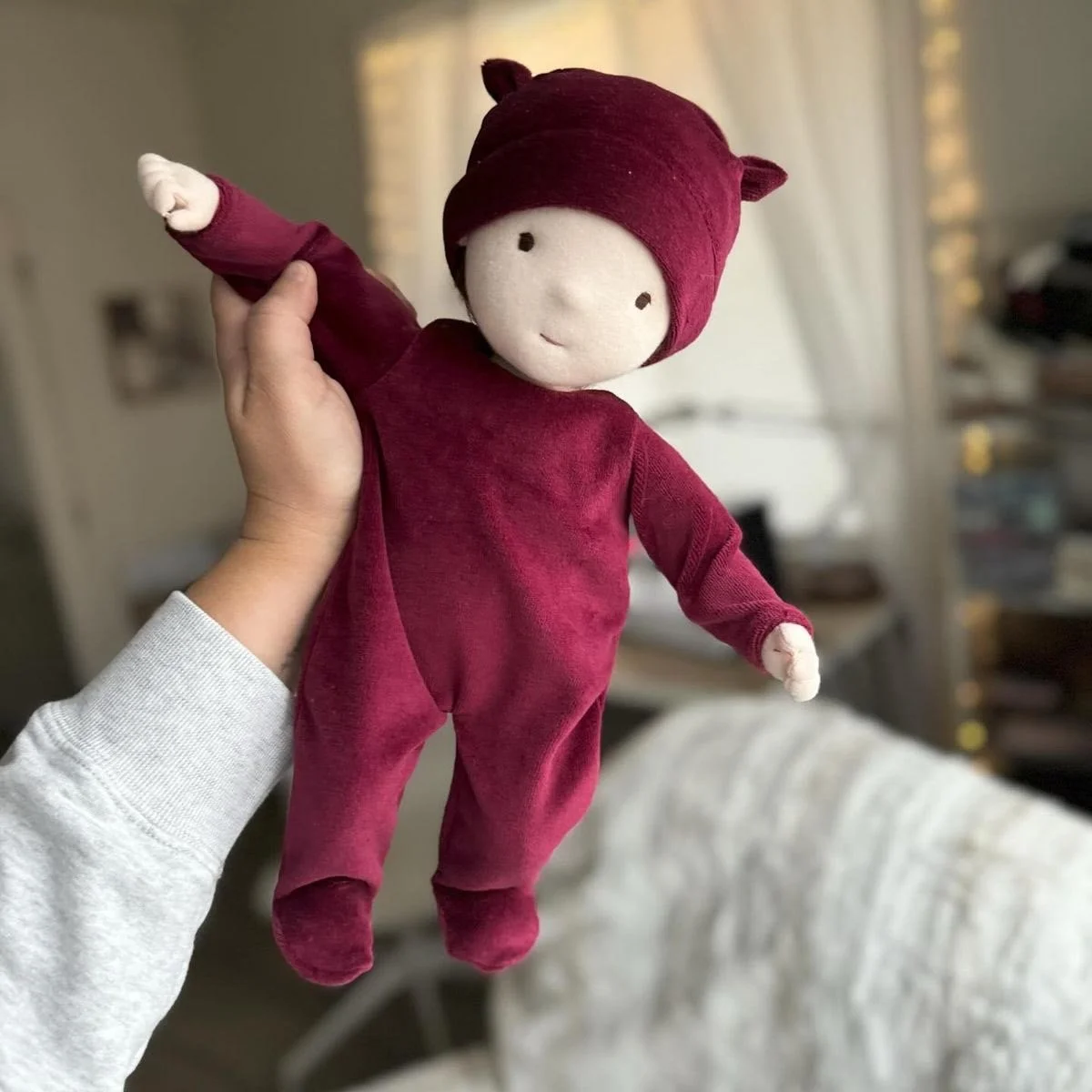 Person holding a soft plush doll wearing a, maroon pajamas and hat with bear ears in a cozy living room.