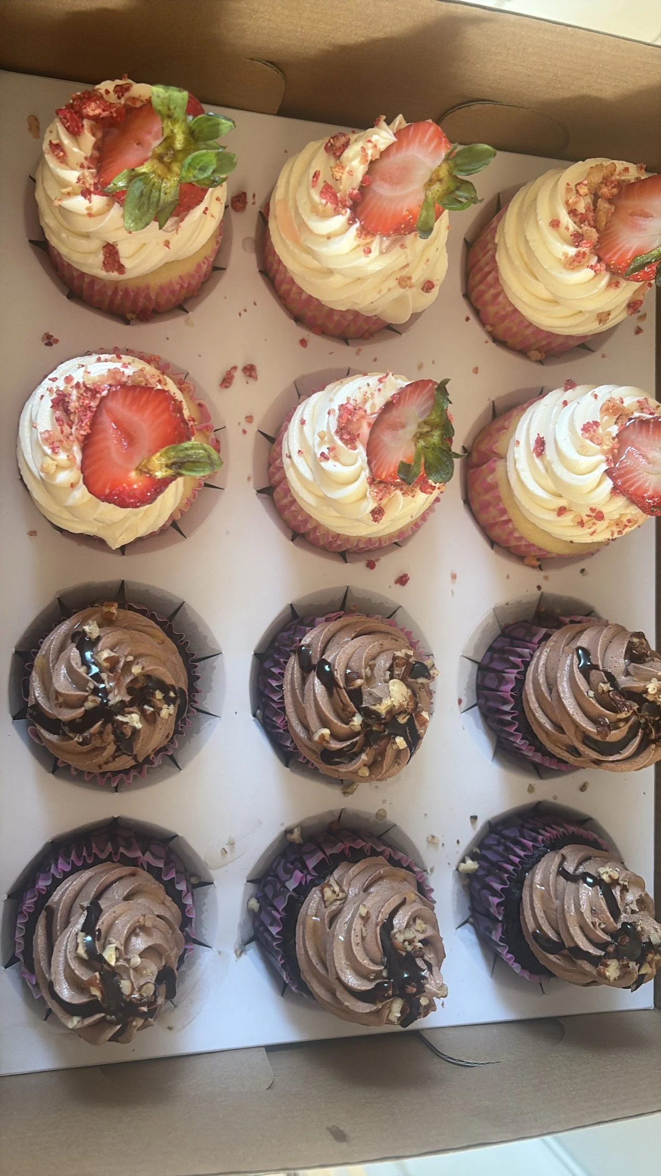 A box of 12 cupcakes, six with strawberries and white frosting, six with chocolate frosting and drizzled with chocolate syrup.