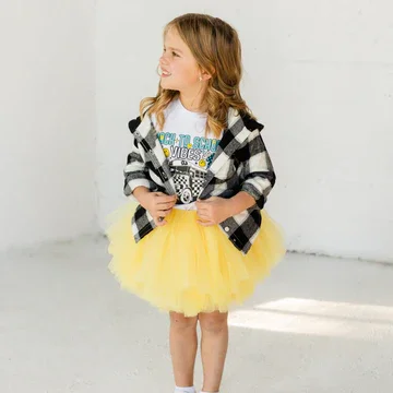 A young girl wearing a yellow tutu skirt, a graphic t-shirt, and a black and white checkered shirt, standing and smiling.