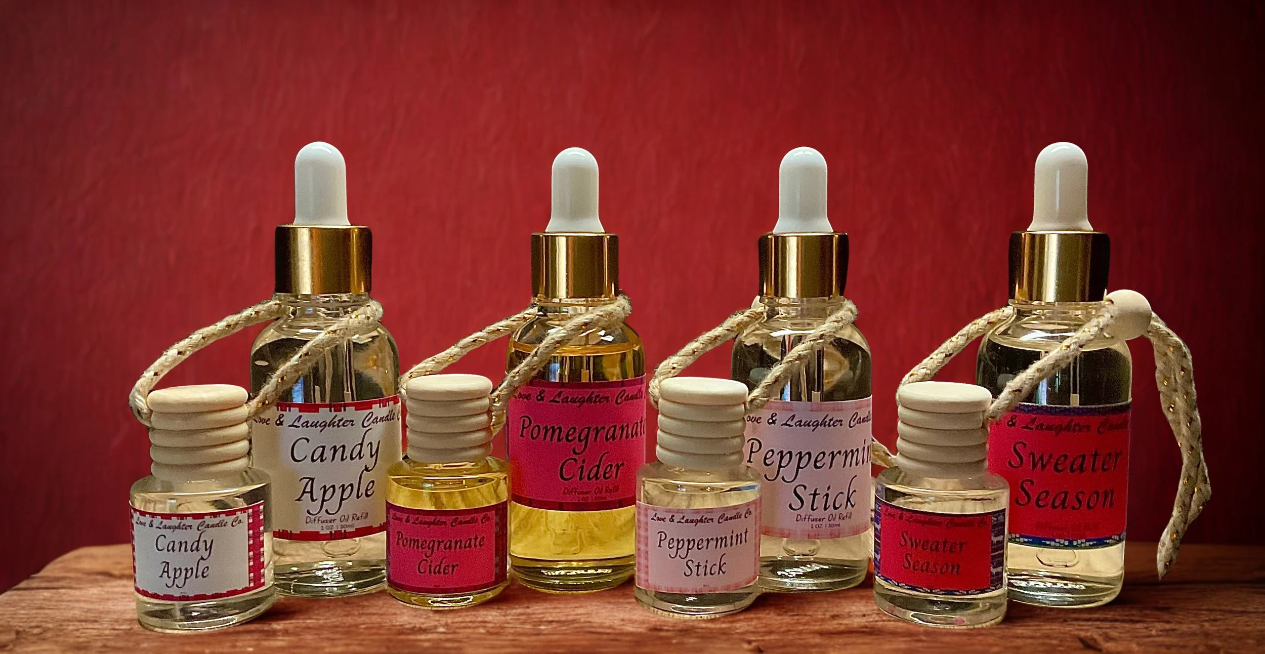 Display of small glass bottles with dropper caps labeled with scents like Candy Apple, Pomegranate Cider, Peppermint Stick, and Sweater Season, arranged on a wooden surface with a red background.