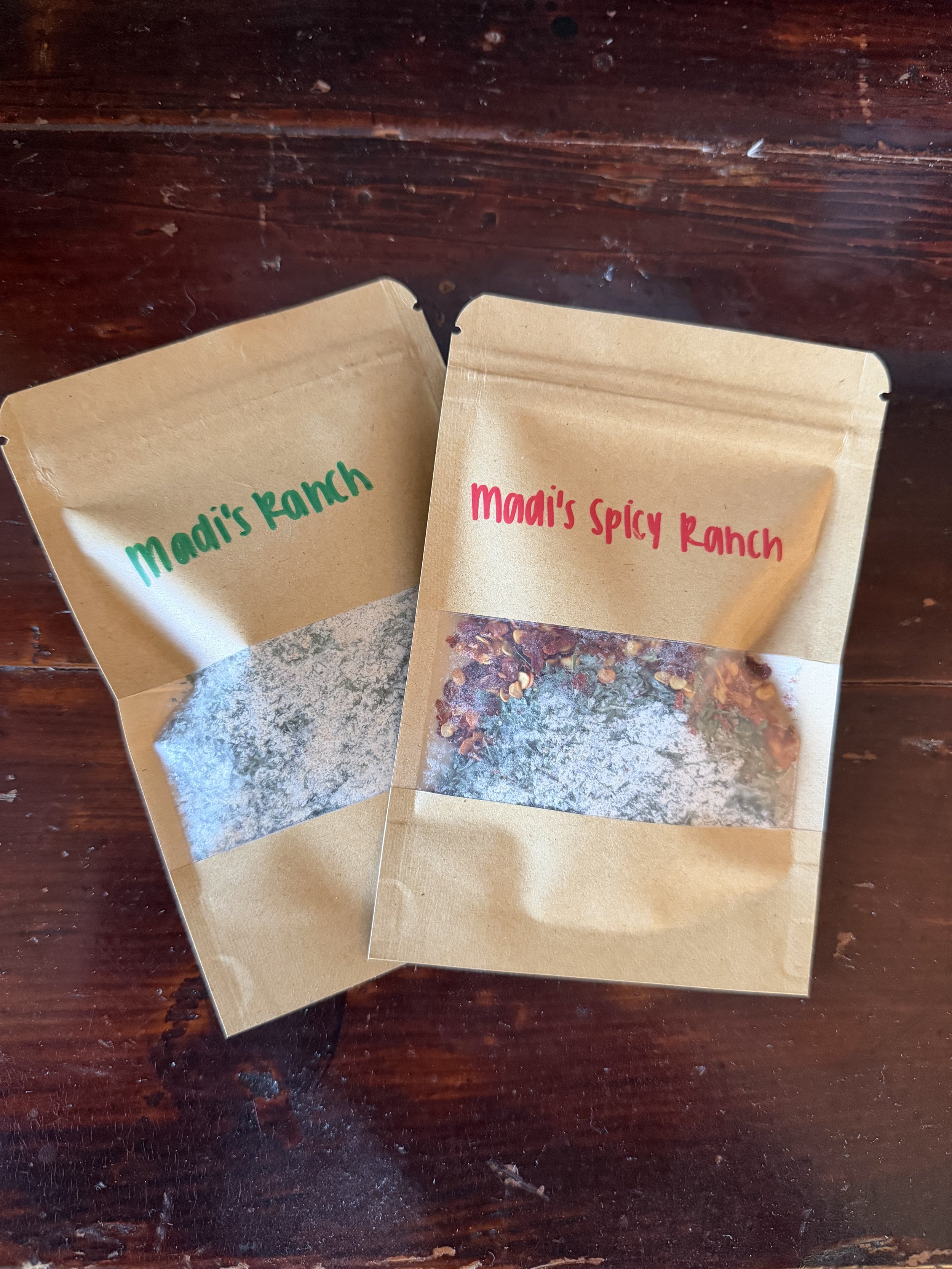 Two brown paper packages labeled 'Madi's Ranch' in green and 'Madi's Spicy Ranch' in red, placed on a dark wooden surface.