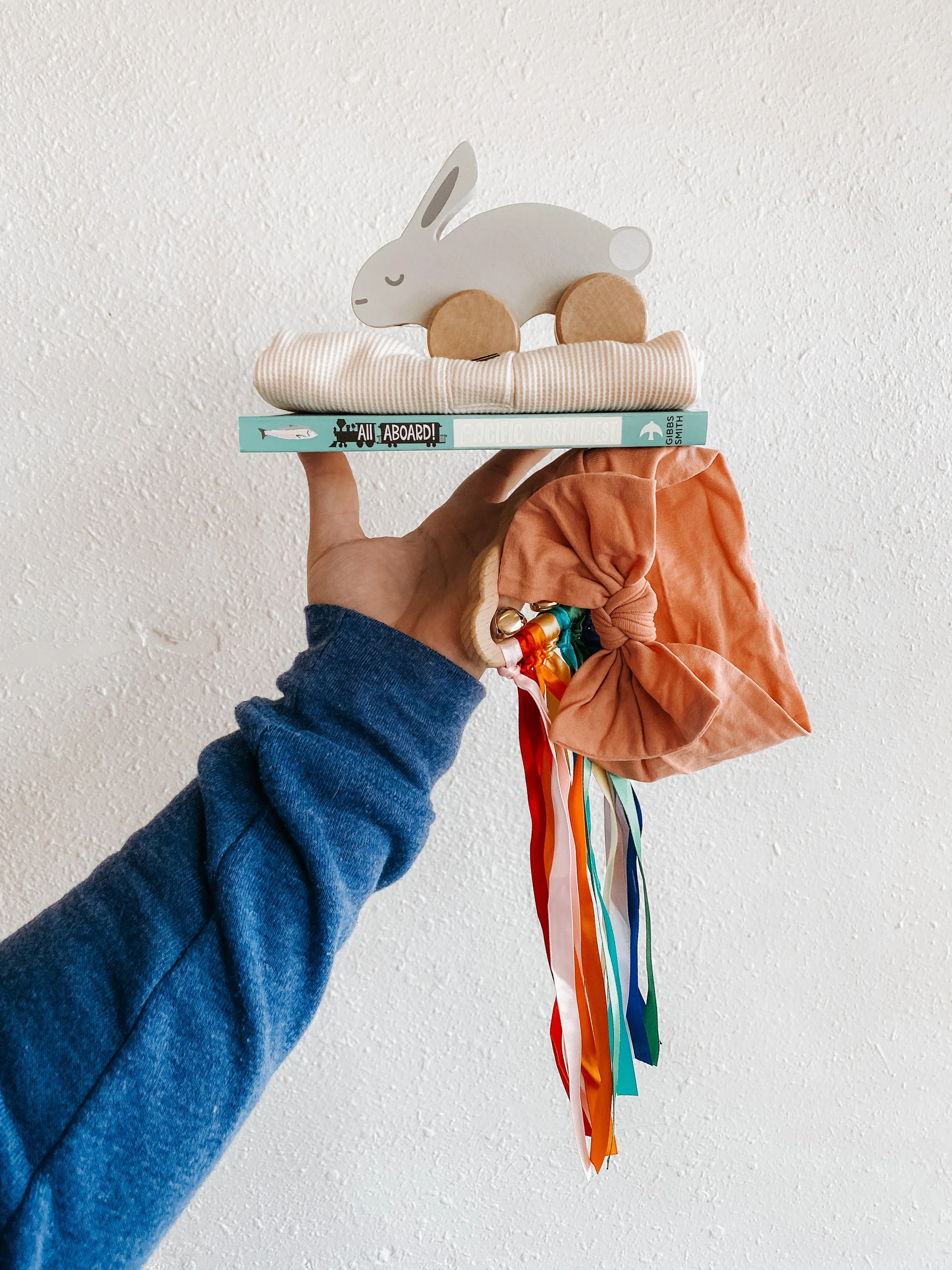 Hand holding a collection of baby items, including a gray plush rabbit toy on wheels, a striped blanket, a children's book titled 'All Aboard!', an orange fabric bag with multicolored ribbons.