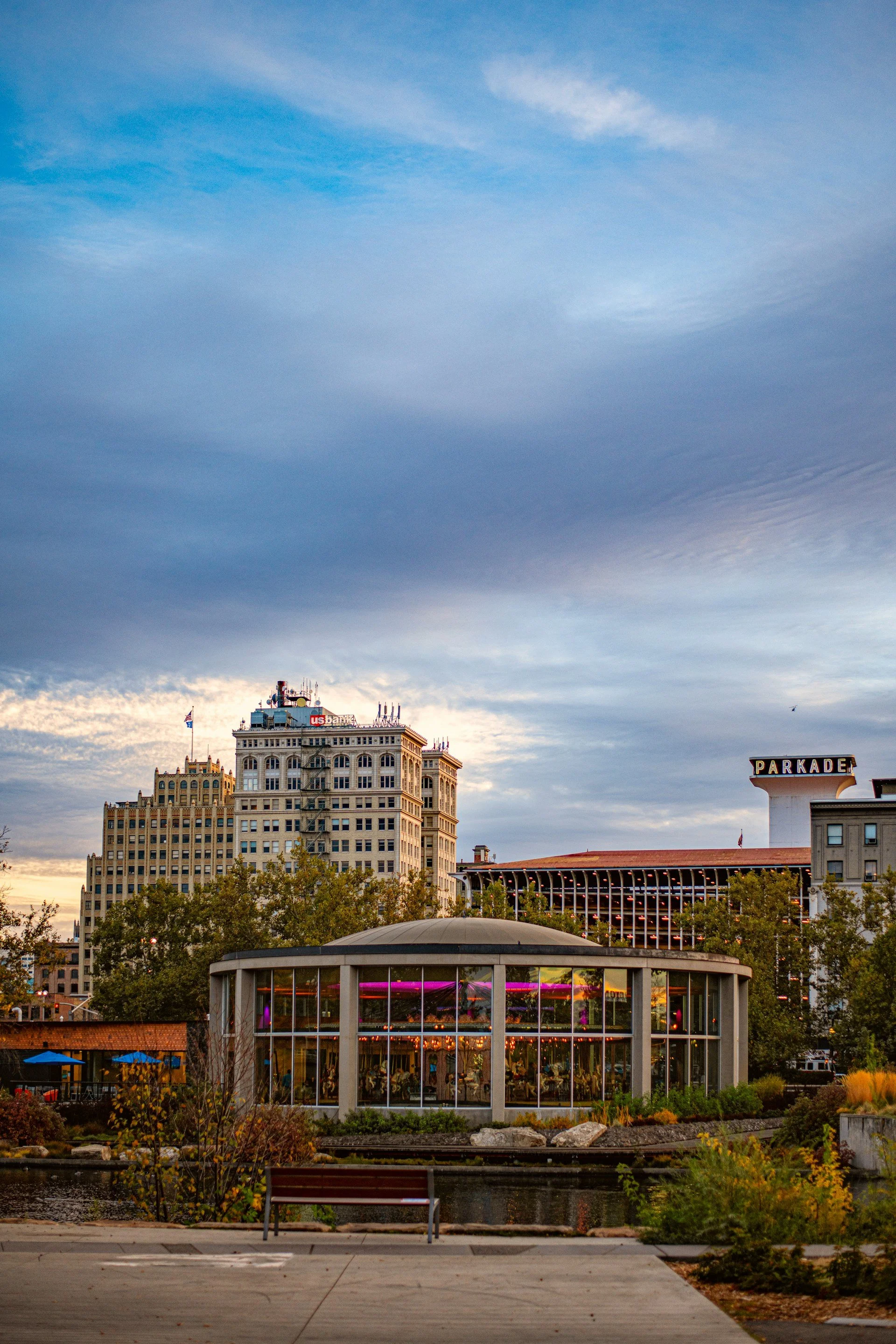 Cityscape with modern buildings and a glass pavilion at sunset, with a blue sky and clouds in the background.