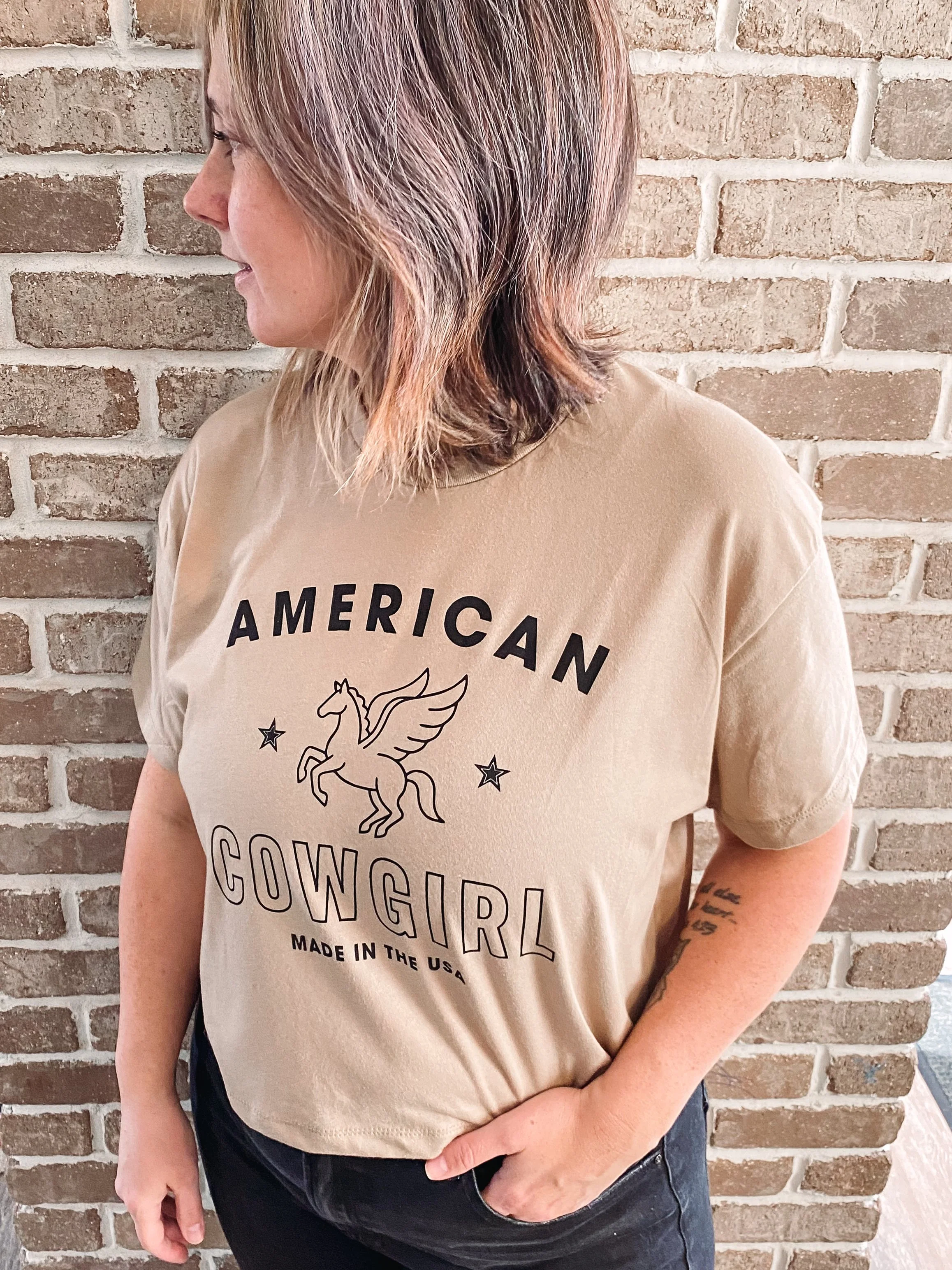 A woman with shoulder-length hair standing in front of a brick wall, wearing a beige T-shirt with the text 'American Cowgirl Made in the USA' and a graphic of a winged horse.