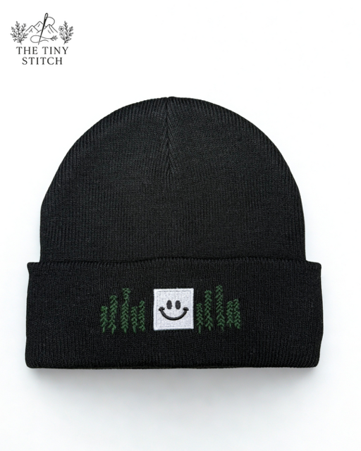 Black knit beanie with a white square patch featuring a smiley face and green embroidered text that appears to spell 'hello' or similar. Logo in top left corner reads 'The Tiny Stitch'.