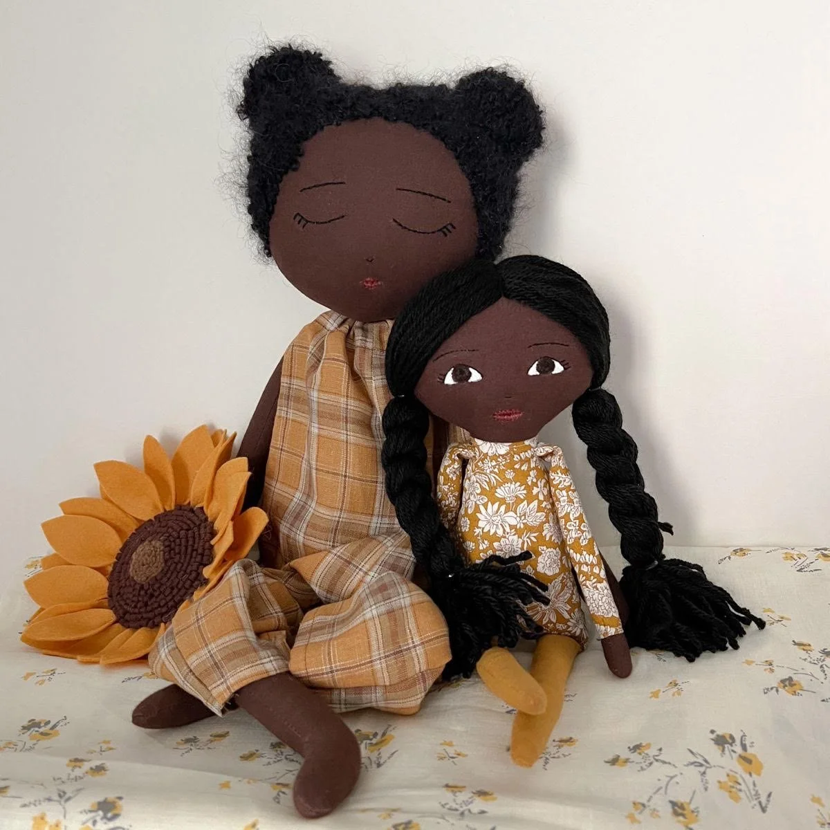 Two handmade cloth dolls, one with closed eyes and curly black hair in a bun, wearing a plaid dress, and the other with open eyes and long black braided hair, wearing a floral dress, sitting on a surface with a sunflower plush toy beside them.