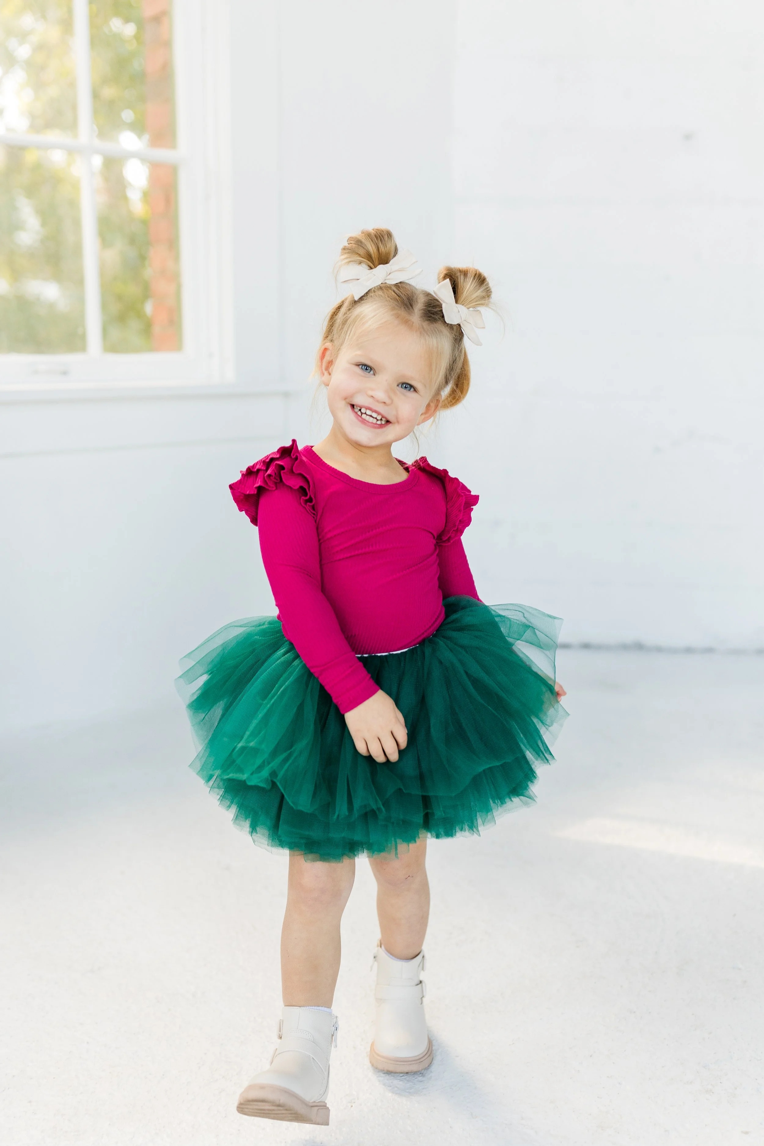 A young girl with blonde hair styled in two buns with bows, smiling and standing in a bright room with large windows, wearing a hot pink long-sleeve shirt, a green tulle tutu, and white boots.