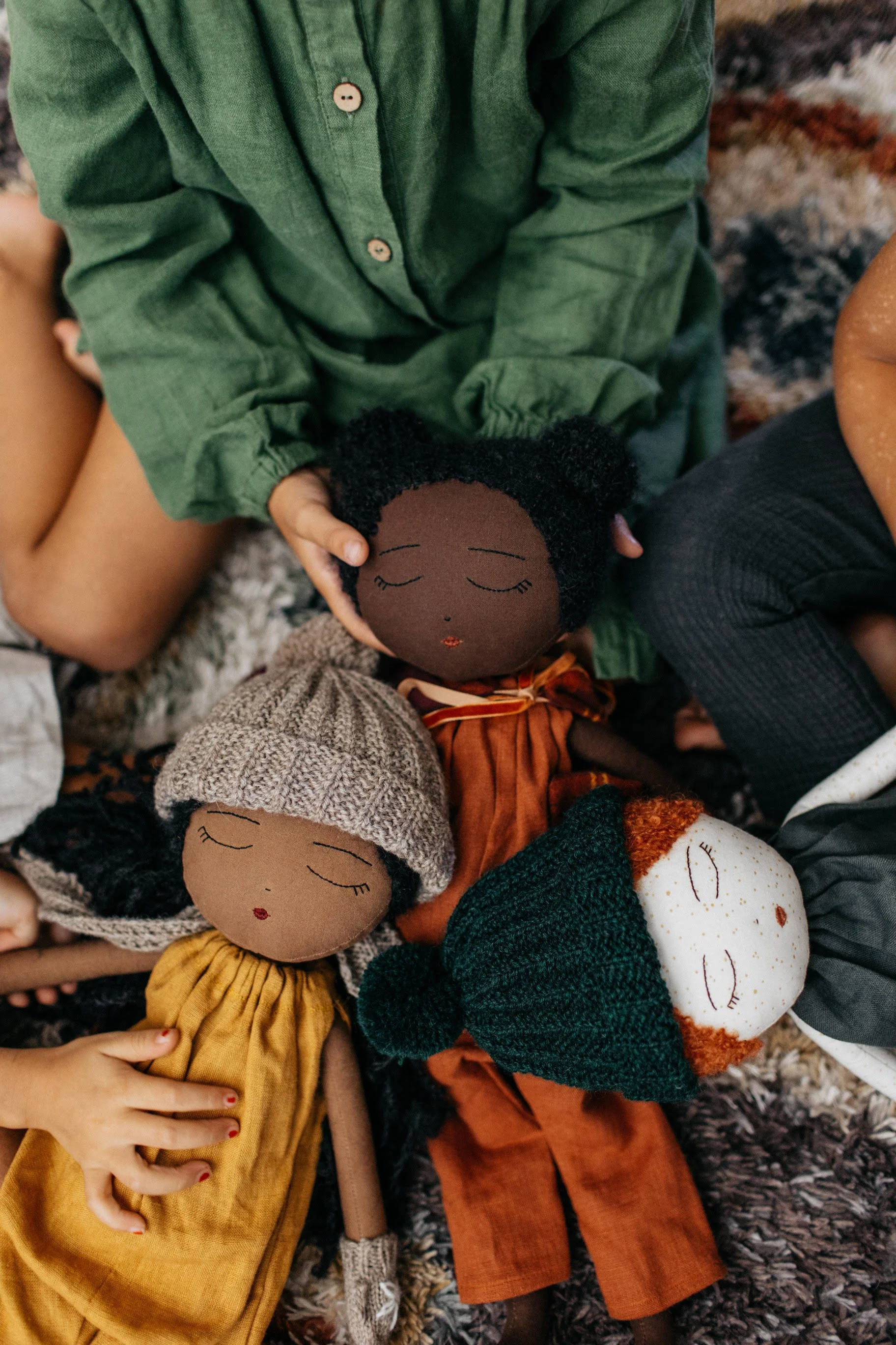 Four handmade cloth dolls resting on a textured rug, with children’s hands gently touching them.