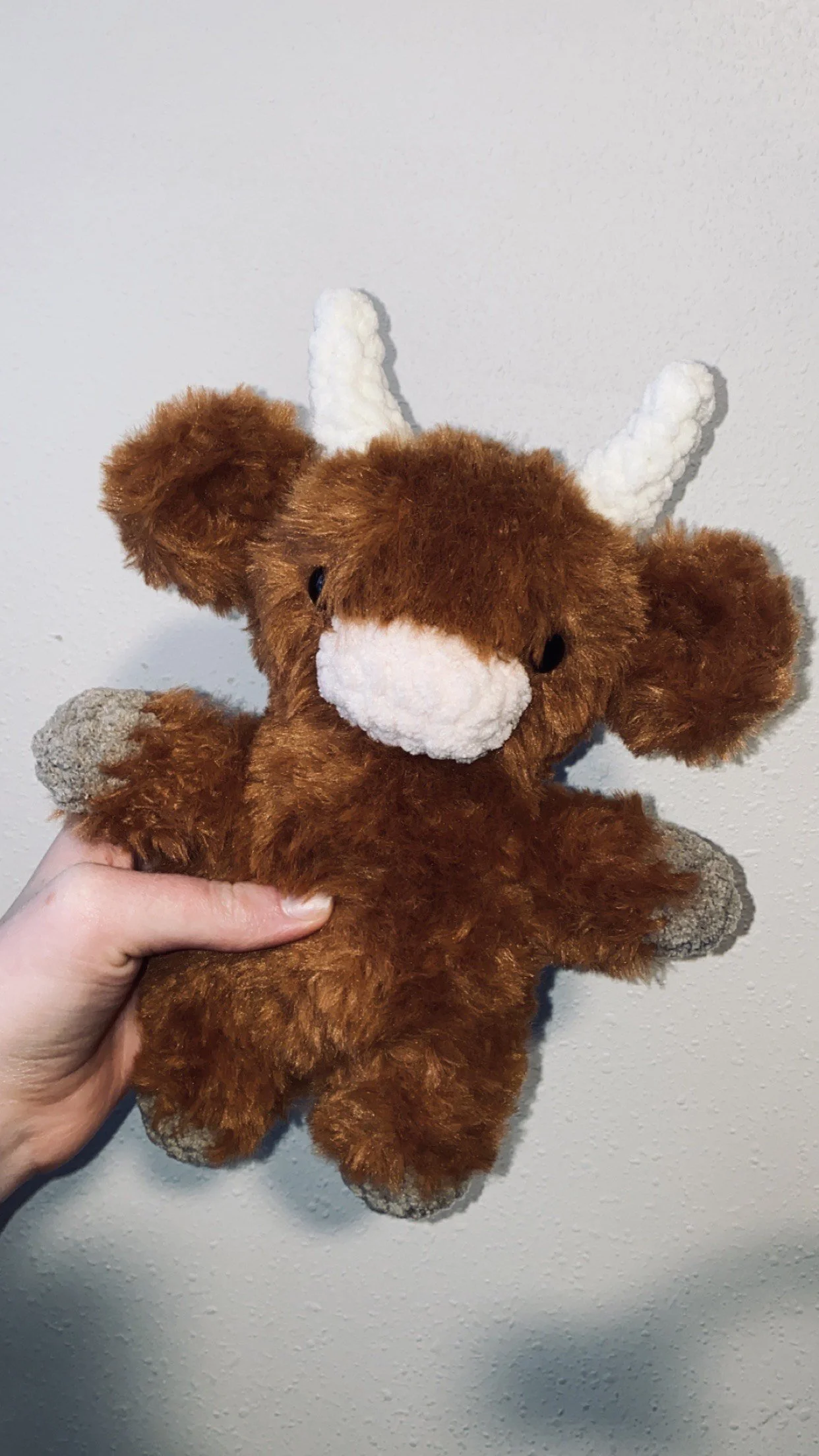 A plush toy teddy bear with white antlers and a white nose being held against a plain wall.