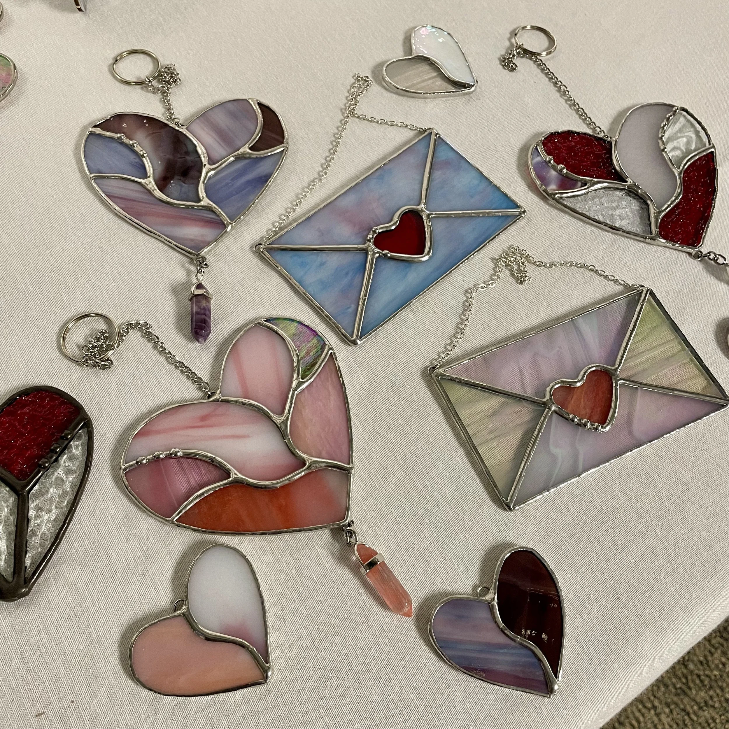 Collection of stained glass jewelry, including heart-shaped pendants and a rectangular pendant with various colored glass panels and metal borders, displayed on a light-colored fabric surface.