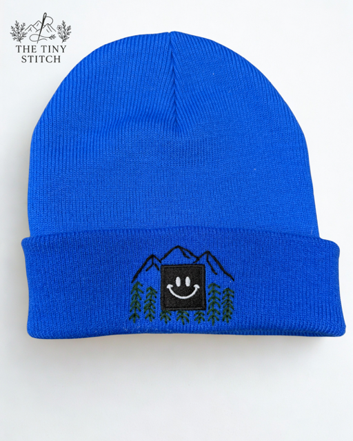Blue knit beanie hat with mountain, tree, and smiley face embroidery.