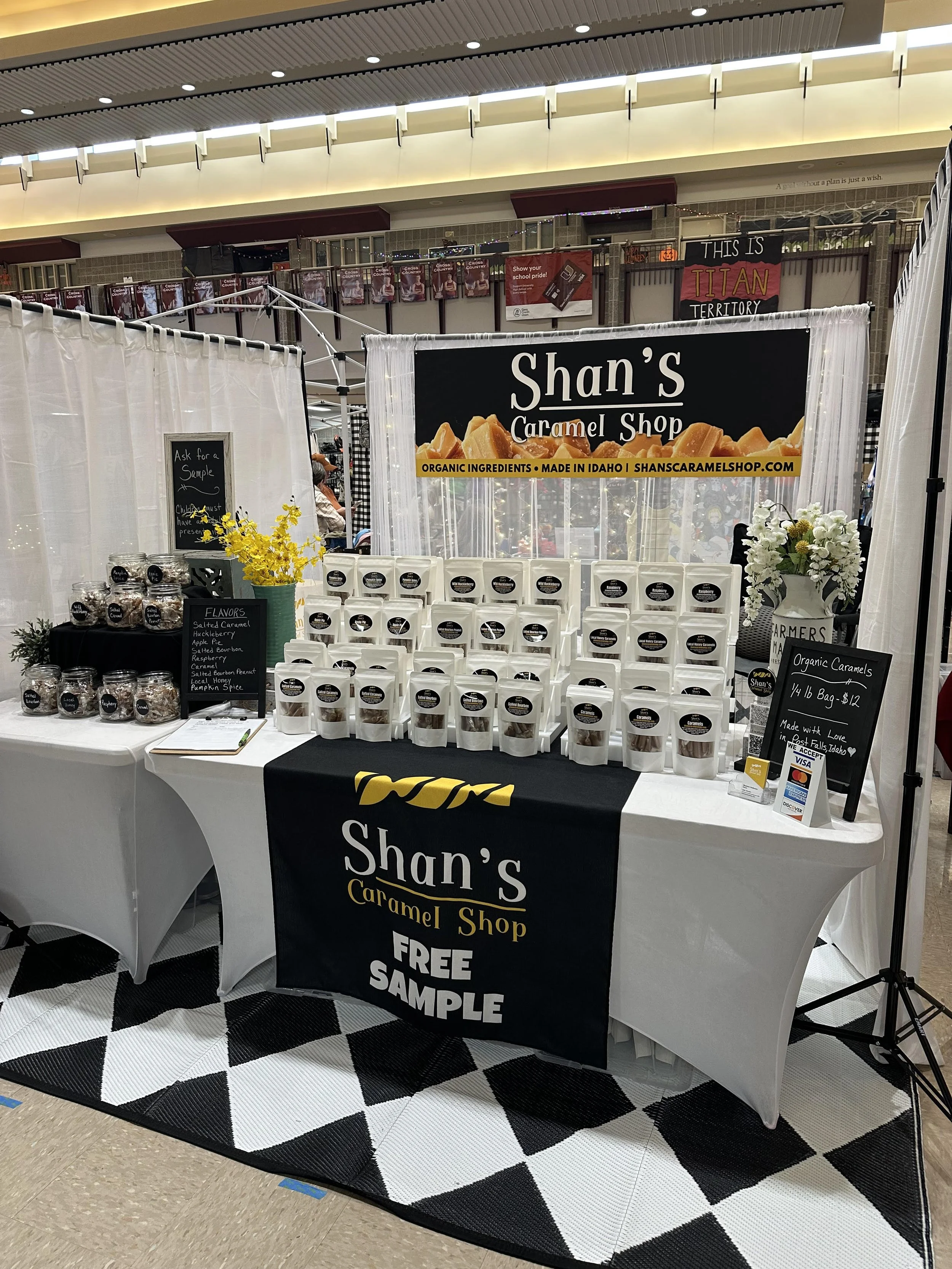 Booth for Shan's Caramel Shop selling organic caramel products, featuring packaged caramel samples on a table with a black and white checkered floor mat, and signs offering free samples.