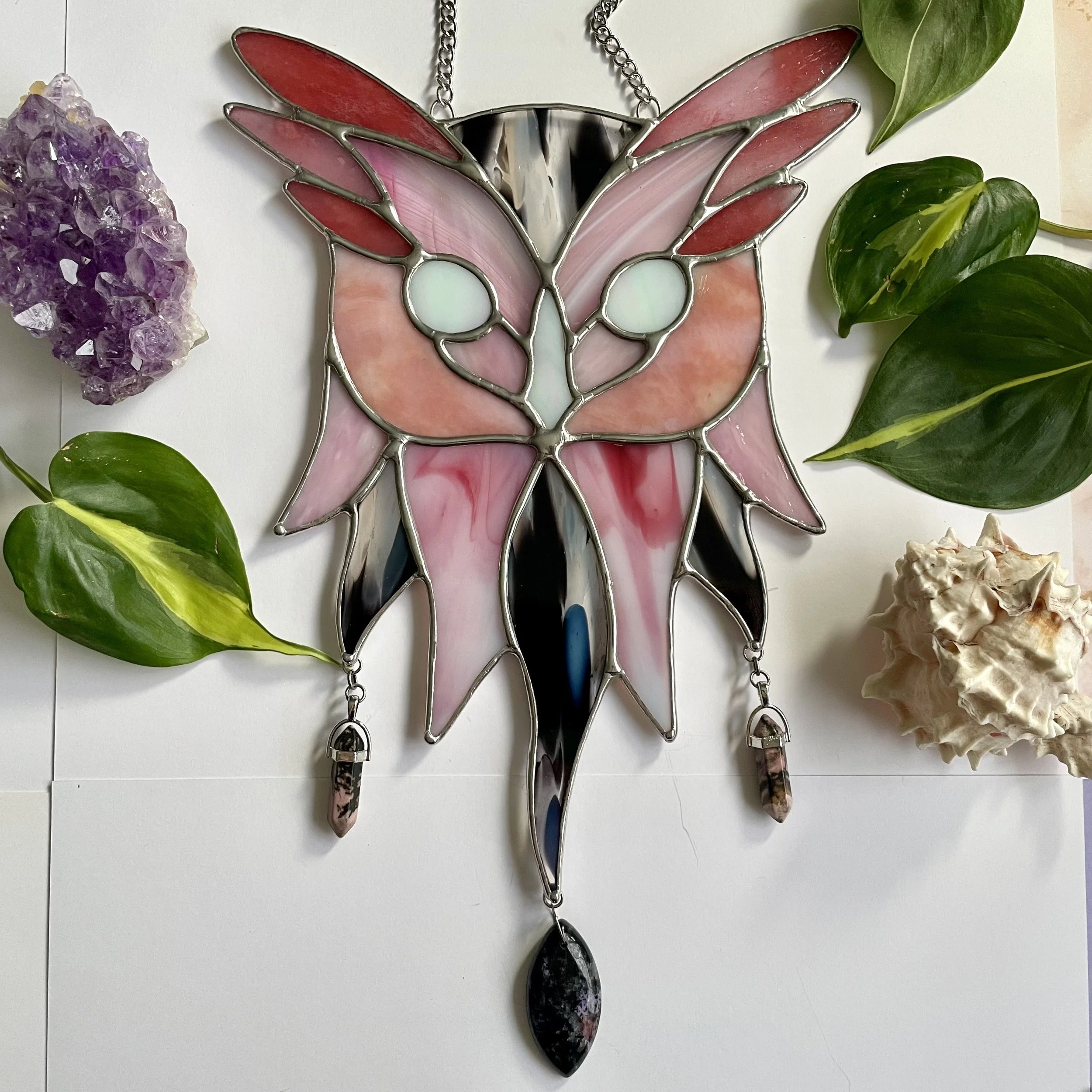 Stained glass art piece in the shape of a butterfly, surrounded by green leaves, a purple crystal cluster, and a seashell.