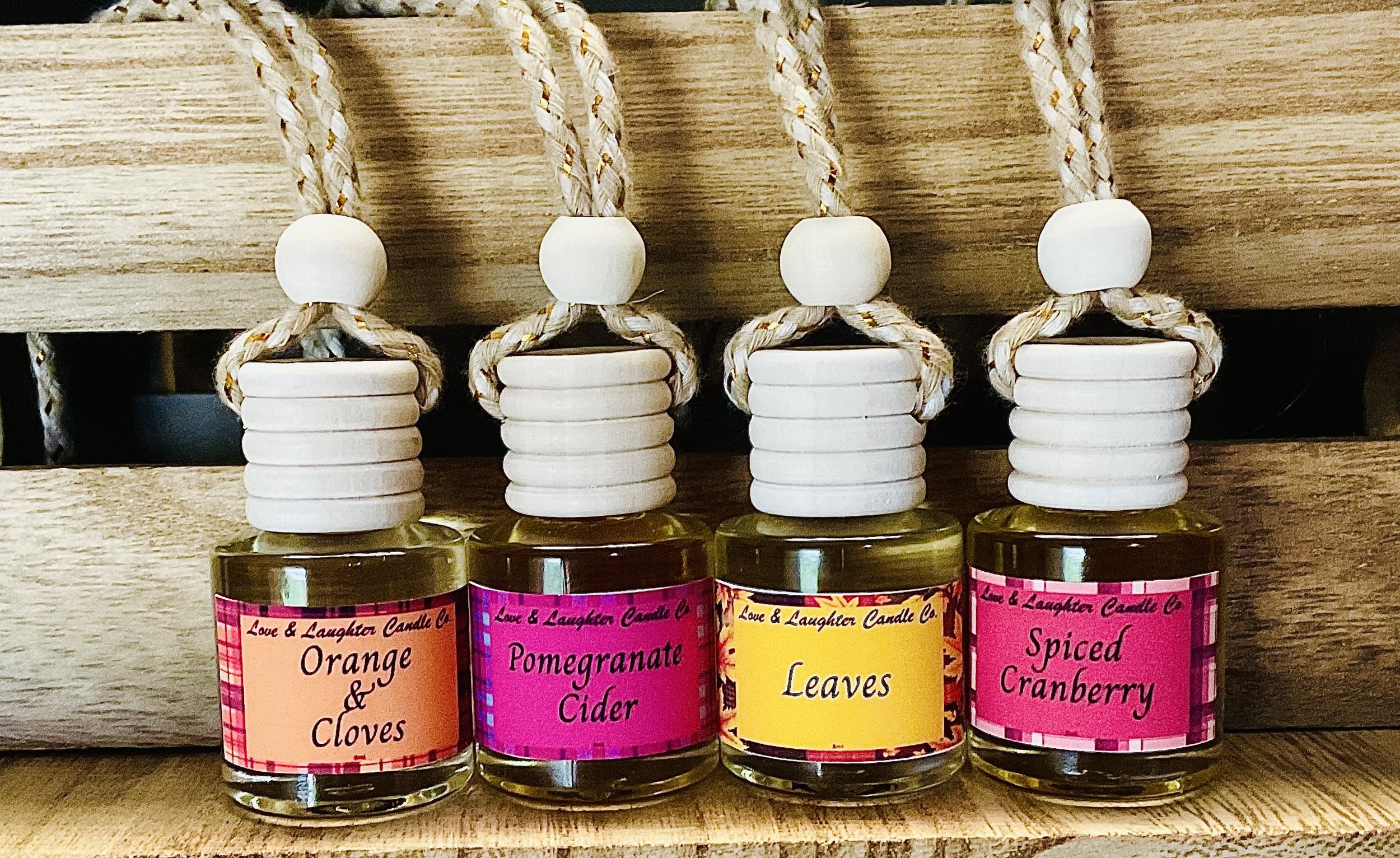 Four candles from Love & Laughter Candle Co, each labeled with different scents: Orange & Cloves, Pomegranate Cider, Leaves, and Spiced Cranberry, with decorative twine and wooden beads on their wicks.