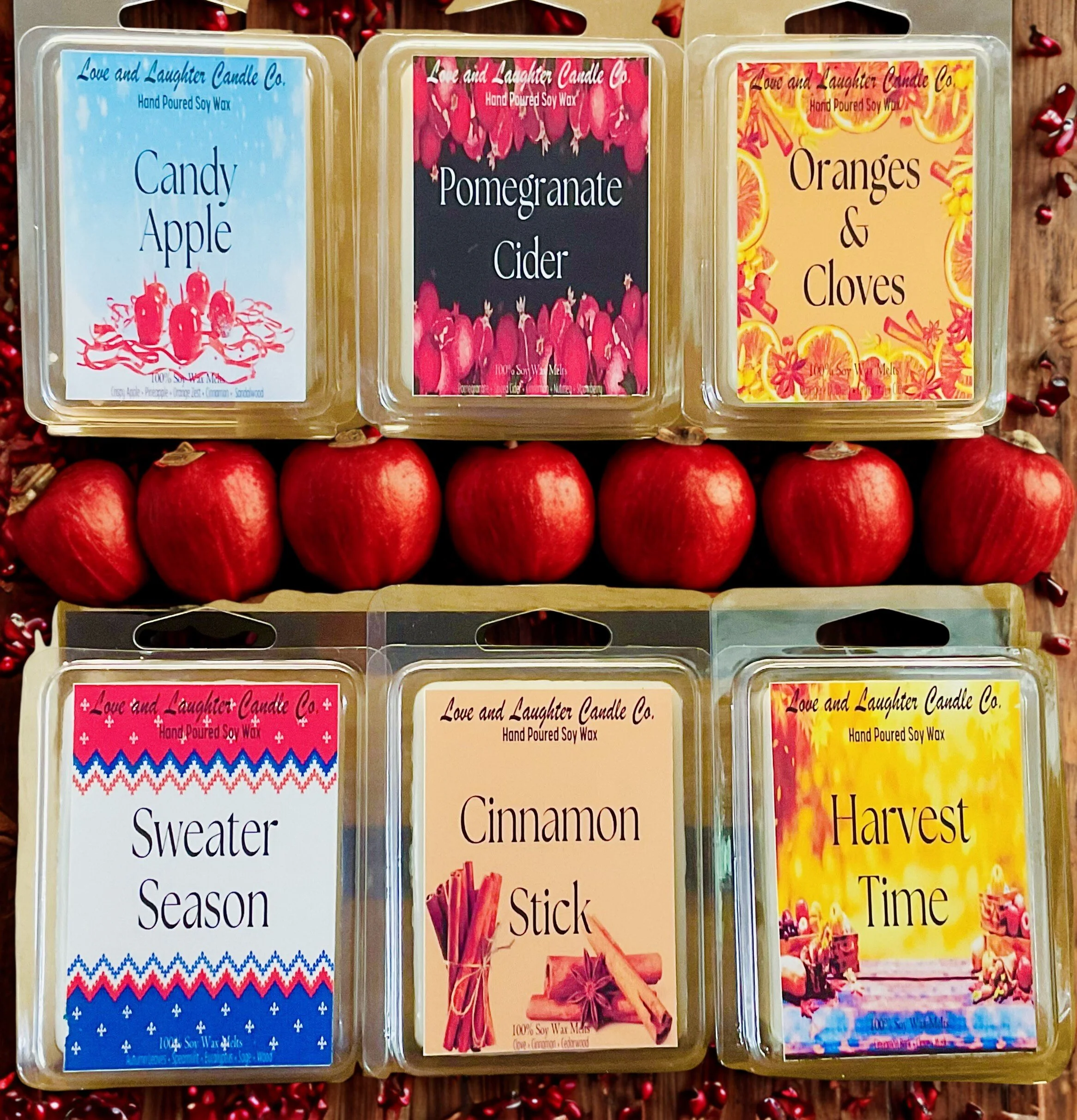 Six scented candles with colorful labels, layered in two rows on a background of red apples and red berries. The top row contains Candyt Apple, Pomegranate Cider, and Oranges & Cloves candles, and the bottom row has Sweater Season, Cinnamon Stick, an