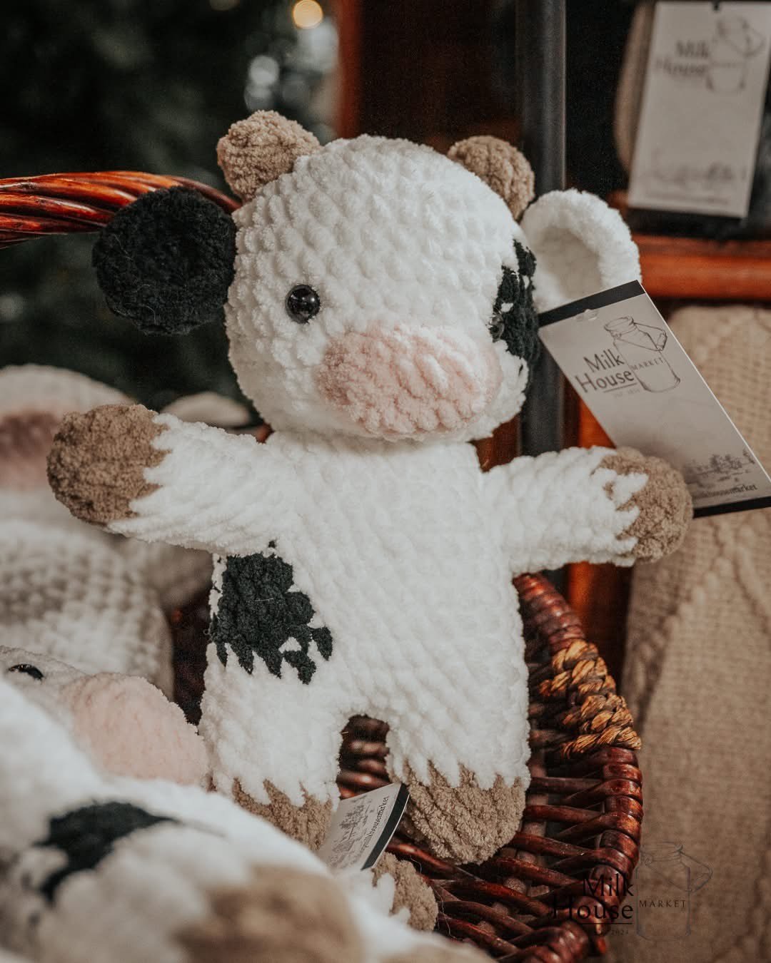 A plush cow toy with a white body, black and pink patches, and brown and black ears, displayed in a basket.