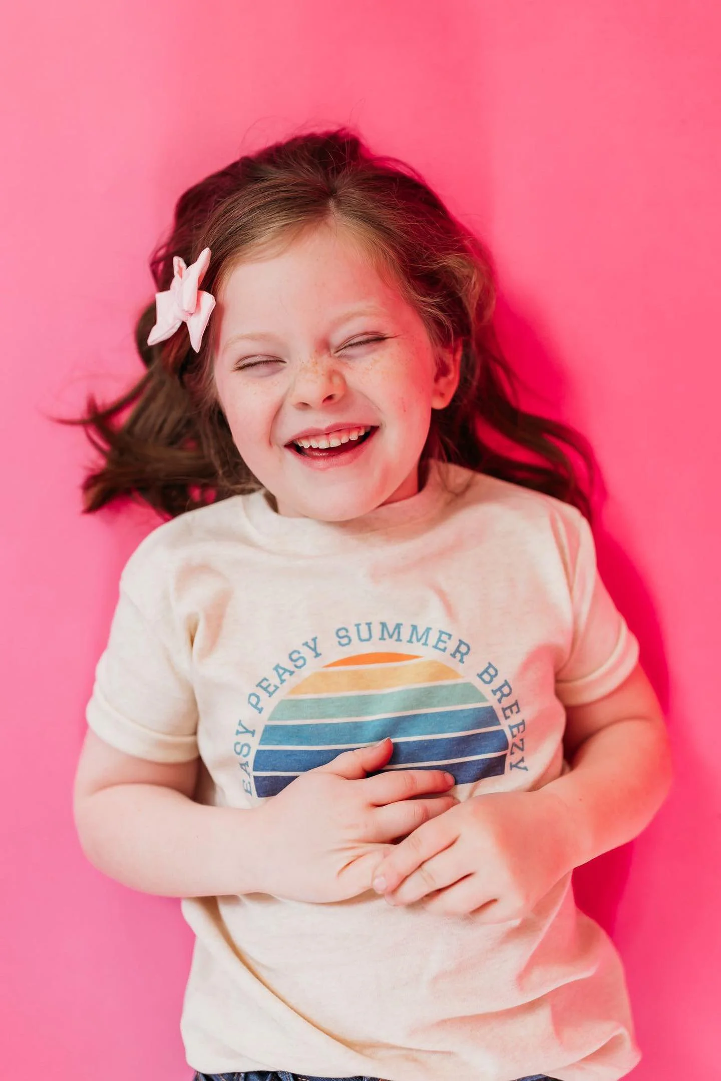 A young girl with long brown hair and a pink bow, lying on a pink surface, smiling and holding her stomach.