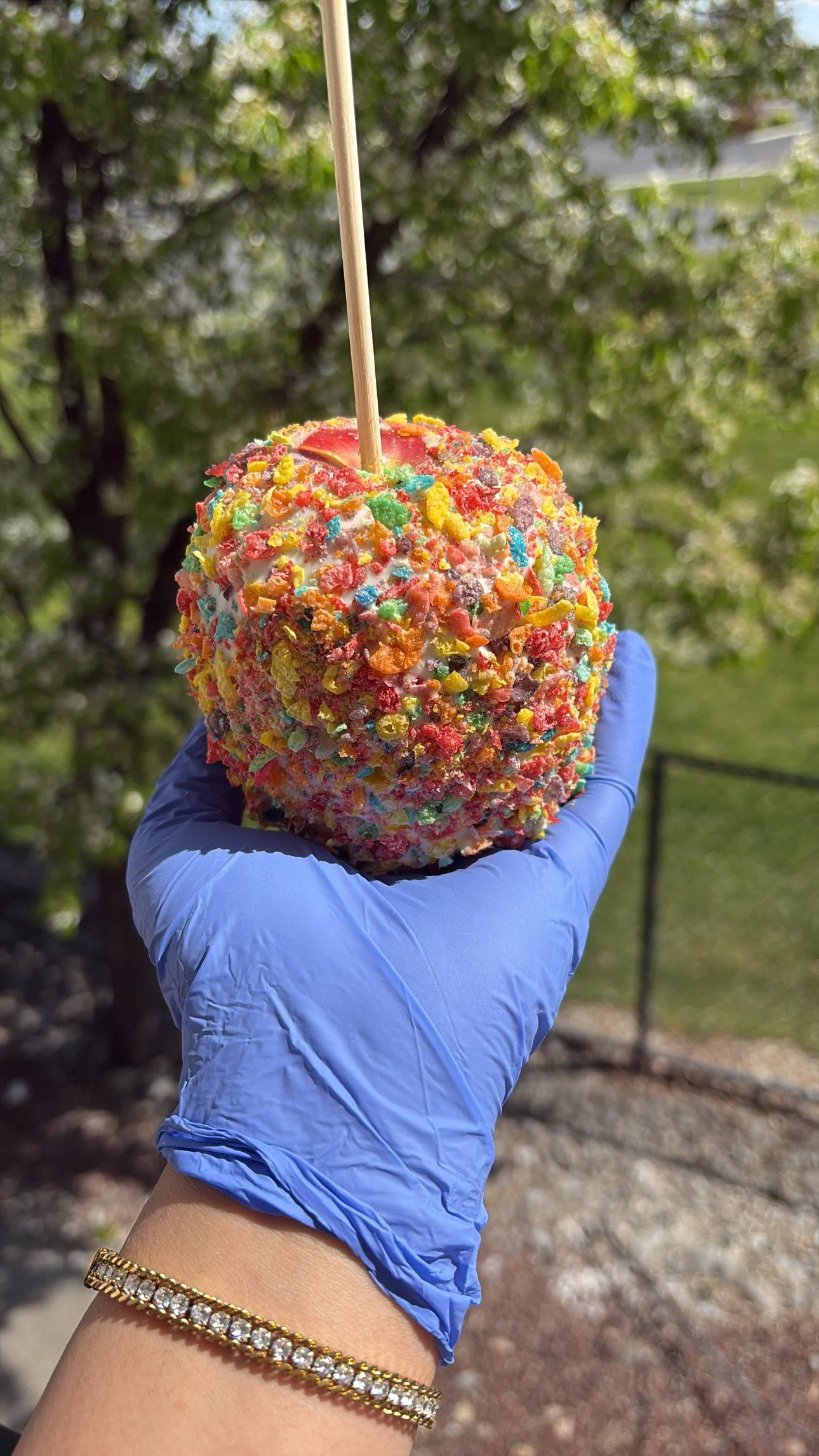A hand wearing a blue glove holding a colorful caramel apple covered in rainbow sprinkles and crushed candies outdoors with trees in the background.