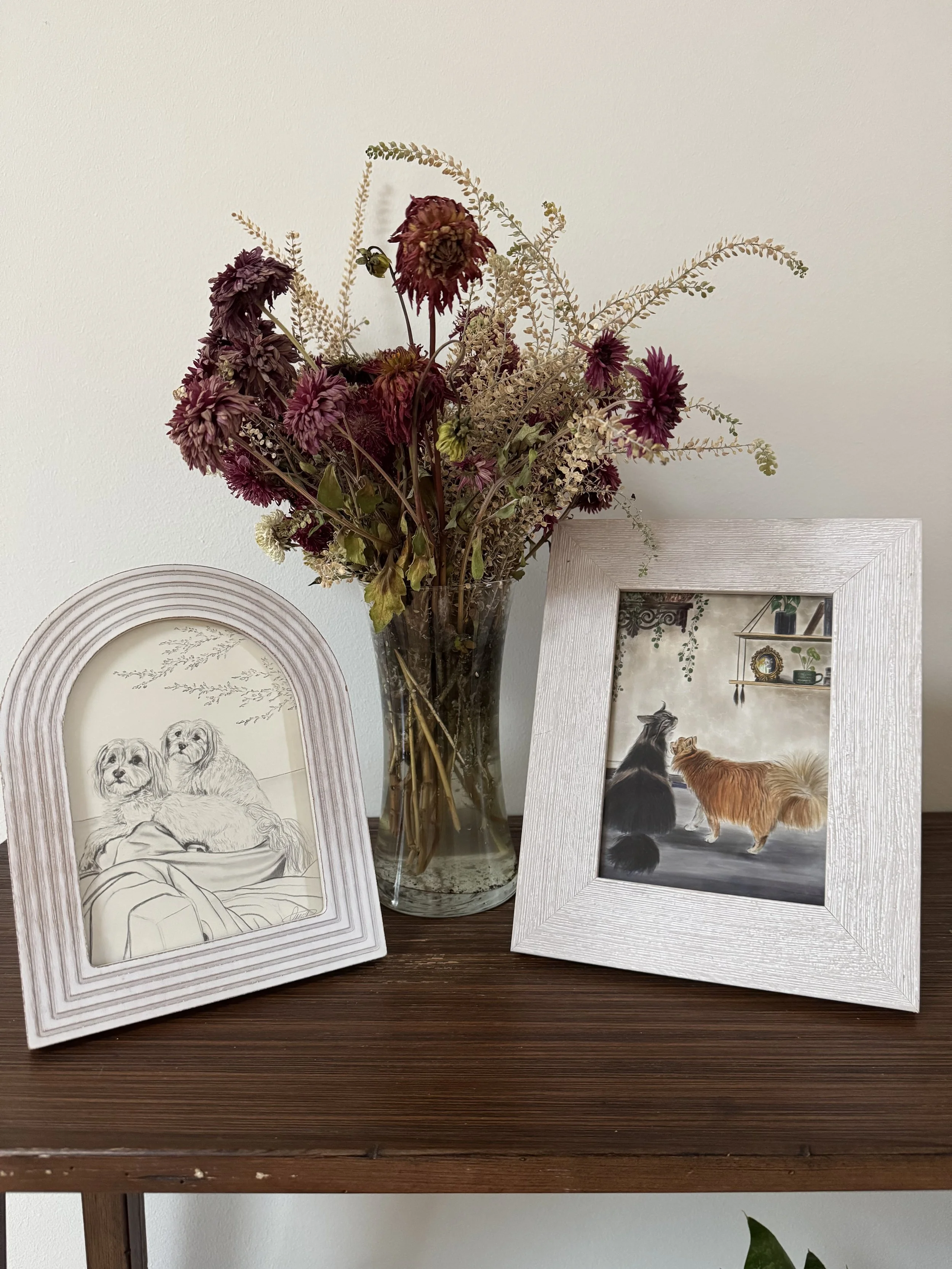 A wooden table holding a clear glass vase with dried purple and beige flowers and two framed pictures of dogs, one being a pencil sketch of two dogs sitting on a blanket, and the other a painting of two dogs, one black and one reddish-brown, in an in