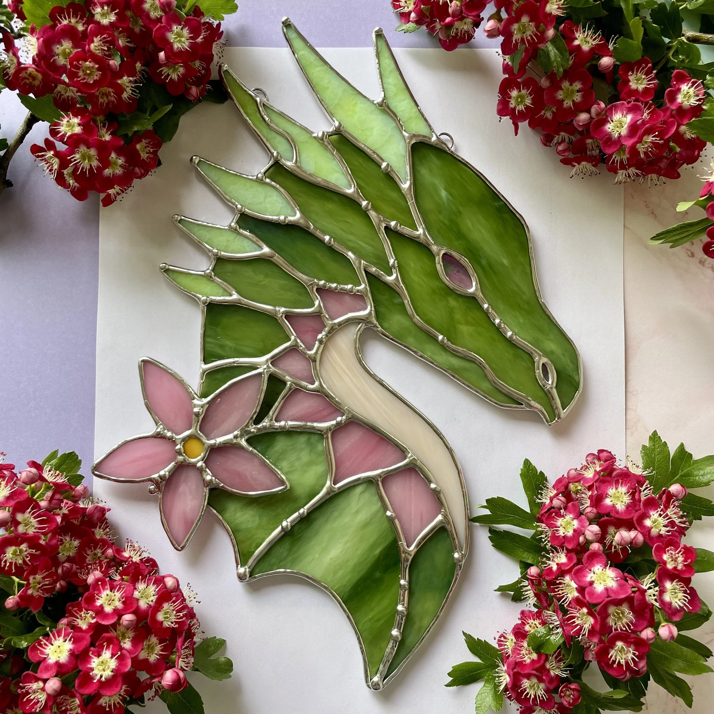 Stained glass art piece of a giraffe with green leaves and pink flowers, surrounded by fresh pink flowers.