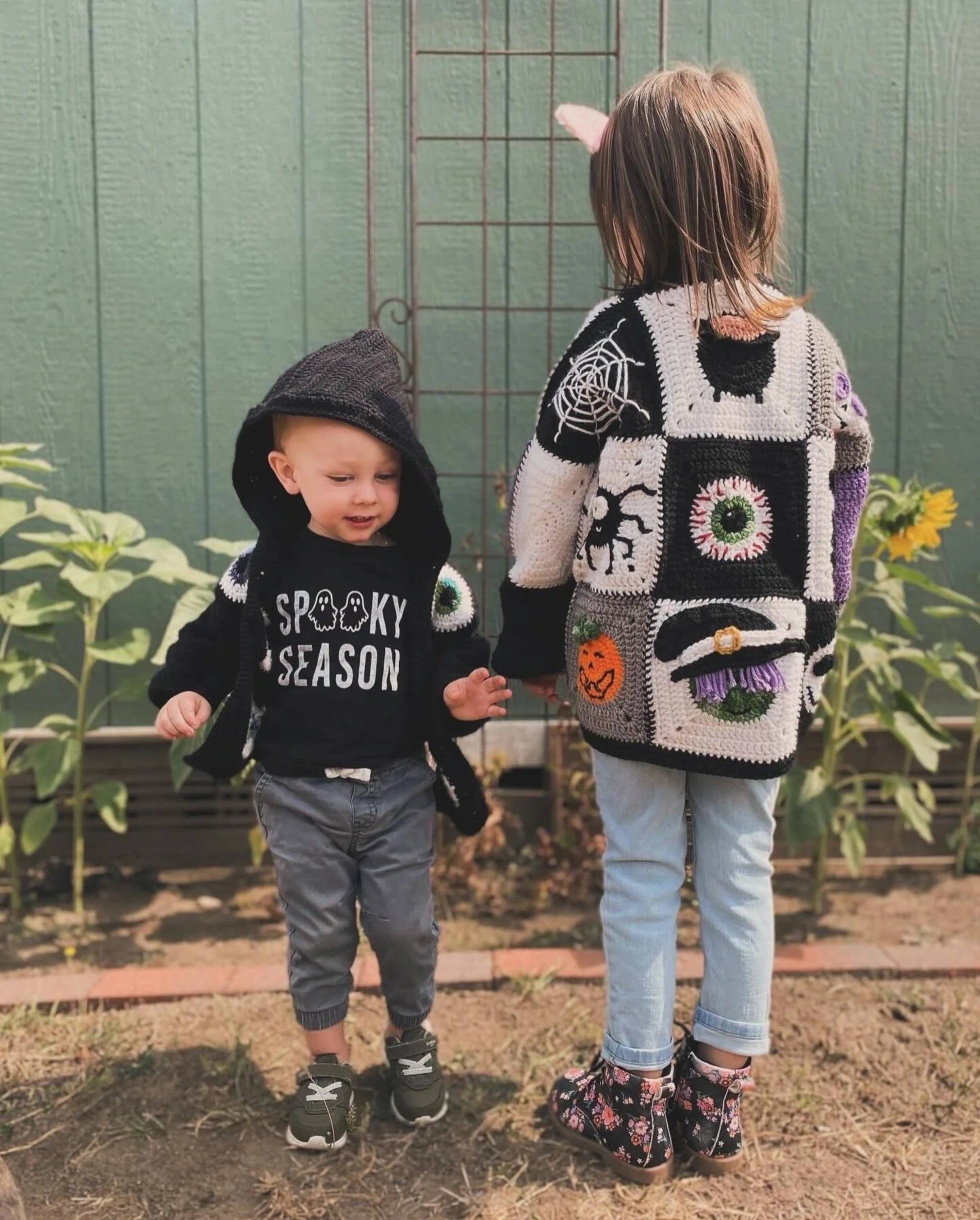 Two children, a younger boy and an older girl, holding hands outdoors on a dirt path in front of a green fence with sunflowers in the background. The boy is wearing a black hoodie with 