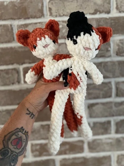Hand holding two yarn dolls, one resembling a fox with orange and white colors and the other resembling a humanoid with black hair and white skin, against a brick wall background.