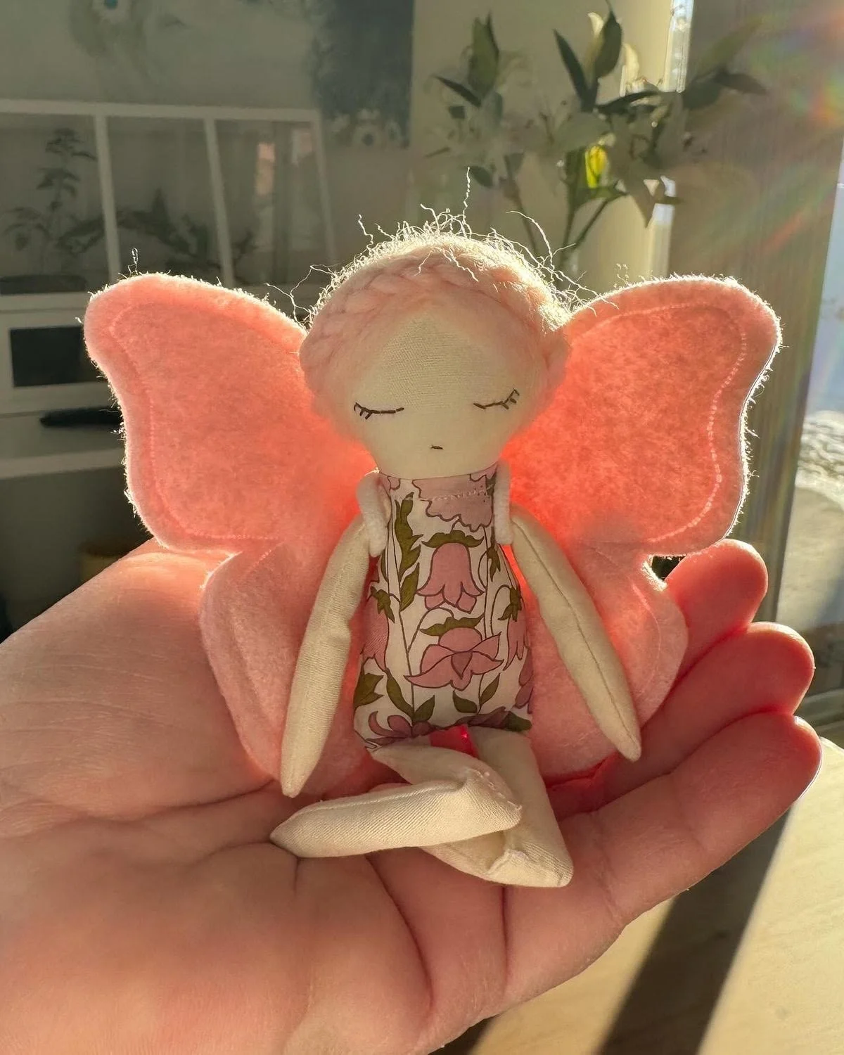 A small doll with pink felt butterfly wings, a floral dress, and long limbs is held in a person's hand. The doll has a simple face with closed eyes and a tiny mouth, and its hair is made of small, light-colored fibers.