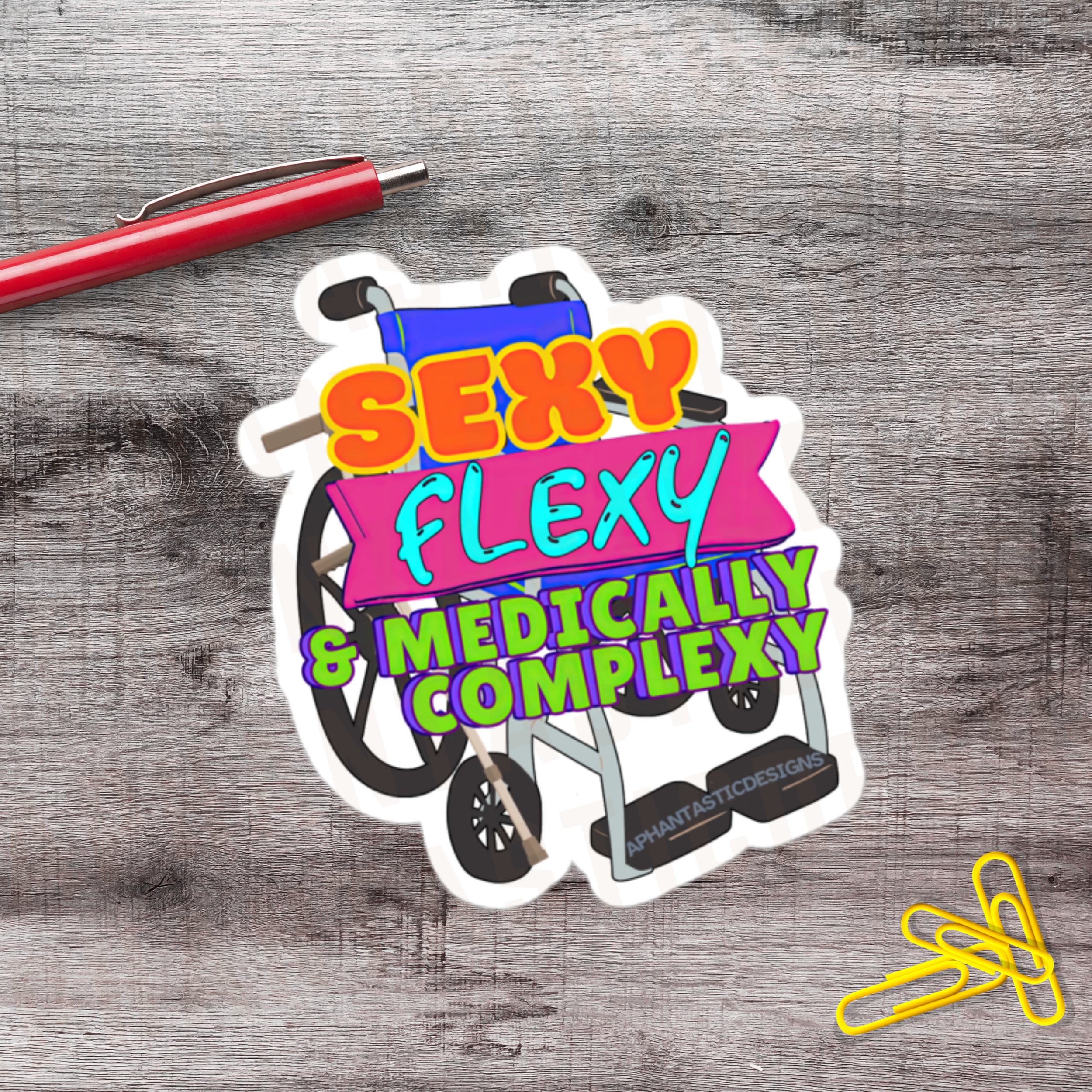 A colorful sticker with text saying "Sexy, Flexy & Medically Complex" next to a red pen and yellow paper clips on a wooden surface.