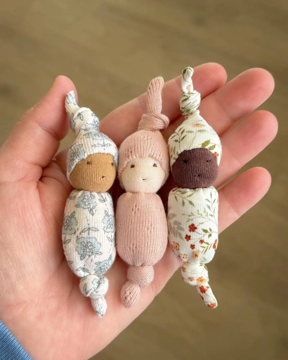 Three small fabric dolls with button bodies and knit heads, each wearing patterned fabric hats, held in a person's hand.