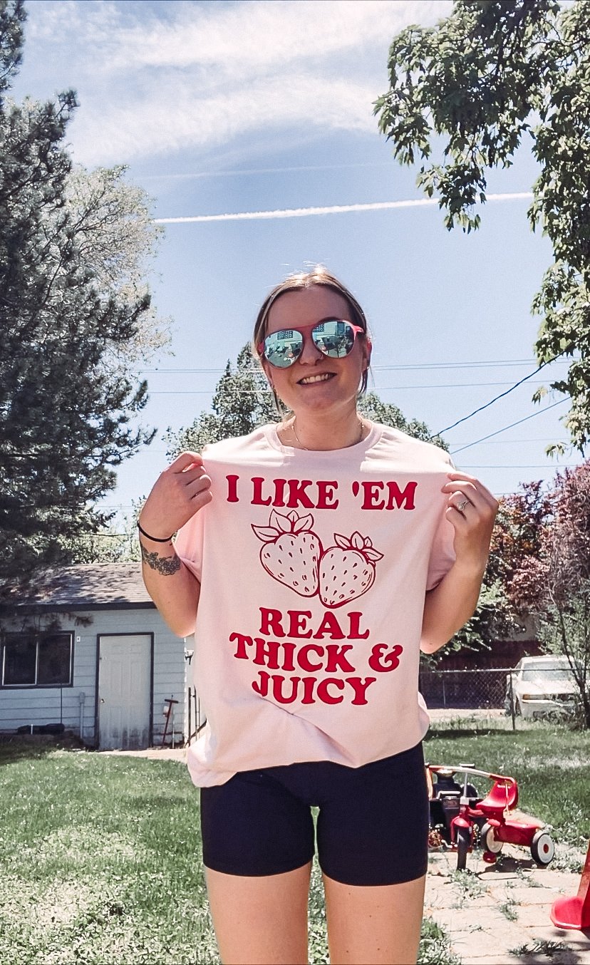 A young woman smiling outdoors on a sunny day, wearing sunglasses and a pink T-shirt with red print and strawberries that says "I LIKE 'EM REAL THICK & JUICY." She is standing on grass with a shed and trees in the background and a small red tricycle 