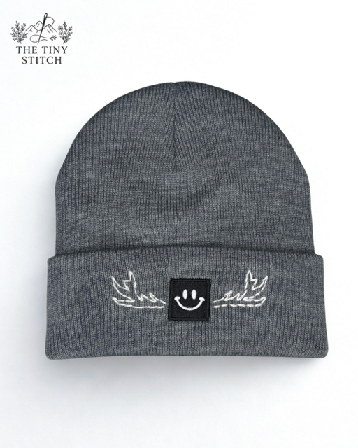 Gray knit beanie hat with a black patch featuring a smiley face and white embroidered wings on the front.