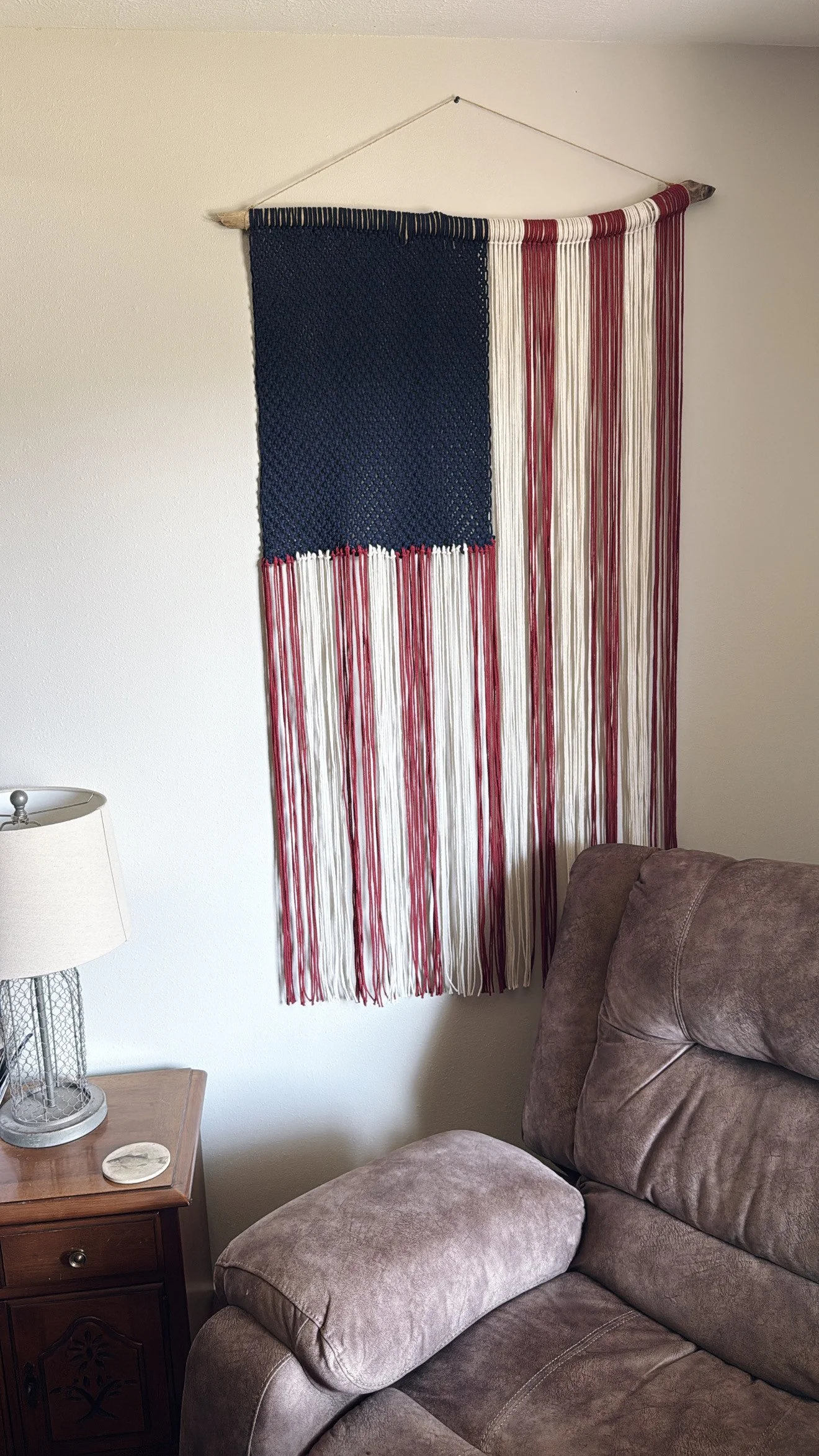 A wall hanging designed to look like the American flag with a blue canton and red and white striped fringe, hung on a wall above a brown plush couch in a living room.