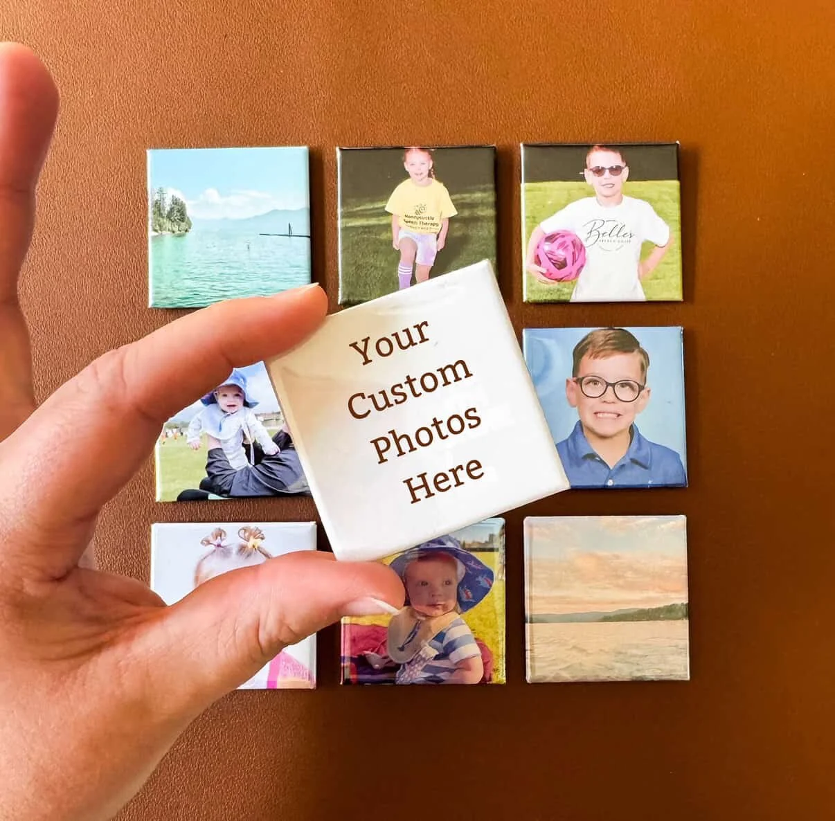 A hand holding a magnet with the text 'Your Custom Photos Here' in front of a background of nine small photo magnets arranged on a brown surface, showing various children and landscapes.