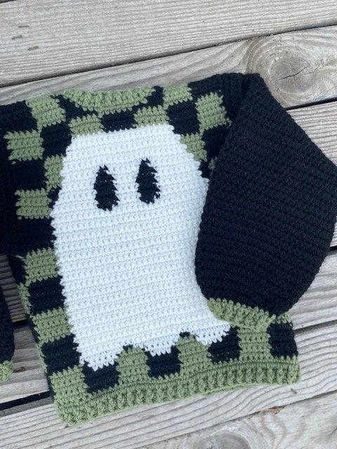 A crocheted Halloween-themed dish towel with a ghost design, green and black border, and a matching black face mask, placed on a wooden surface.