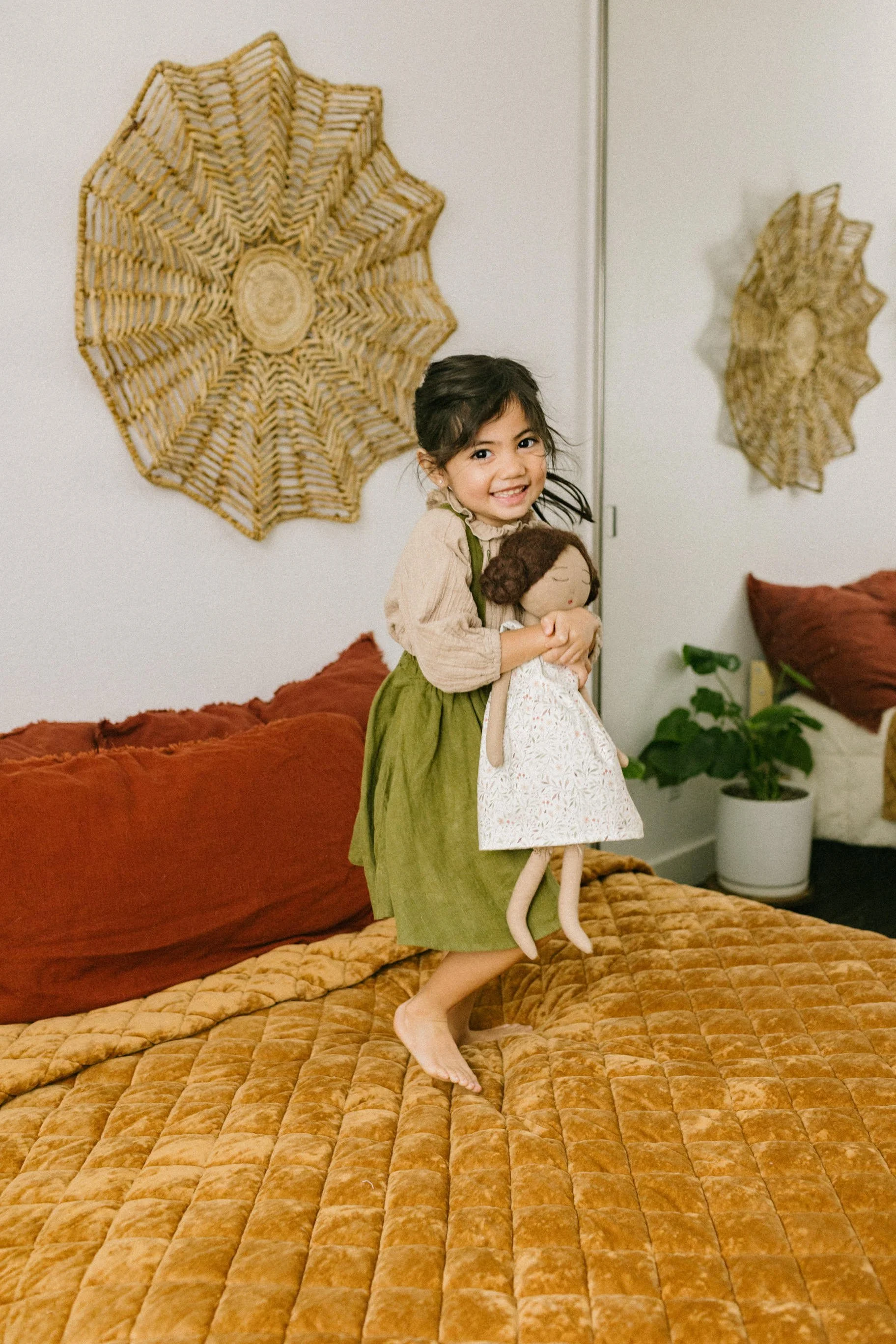A young girl smiling and jumping on a bed while hugging a handmade doll. The room has a woven wicker wall decoration, a plant in a white pot, and warm-toned bedding.