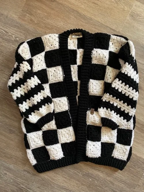 Black and white checkered crocheted sweater or cardigan laid flat on wooden floor.
