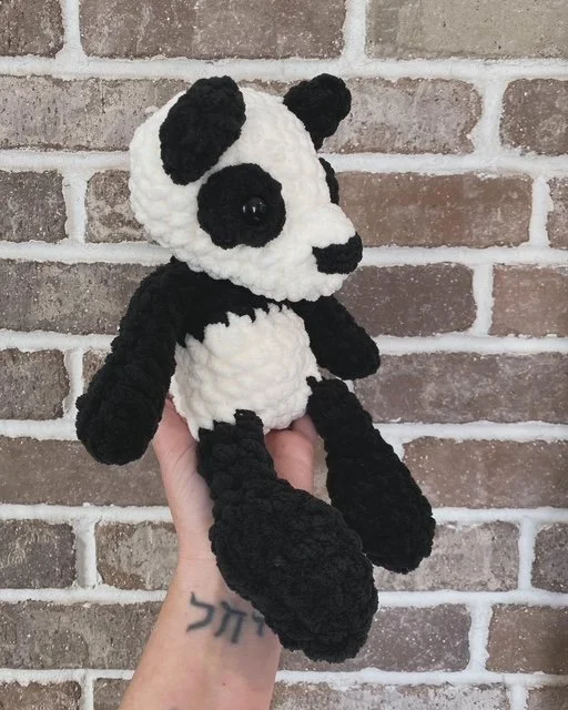 Hand holding a black and white knitted panda stuffed animal against a brick wall background.