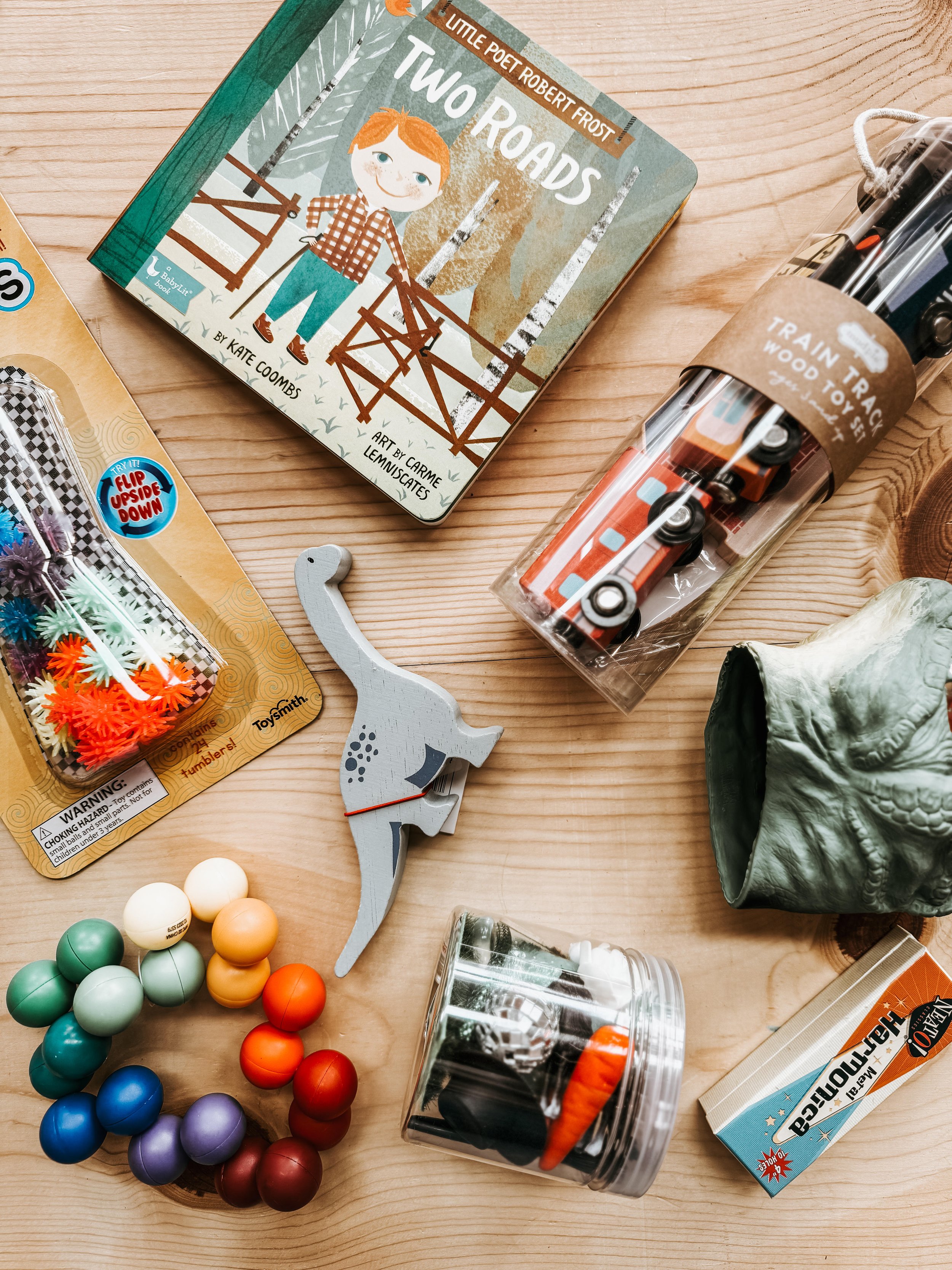Children's toys and books on a wooden surface, including a book titled 'Two Roads' with an illustration of a boy, a train toy set, a dinosaur-shaped toy, a container of colorful balls, and various other small toys.
