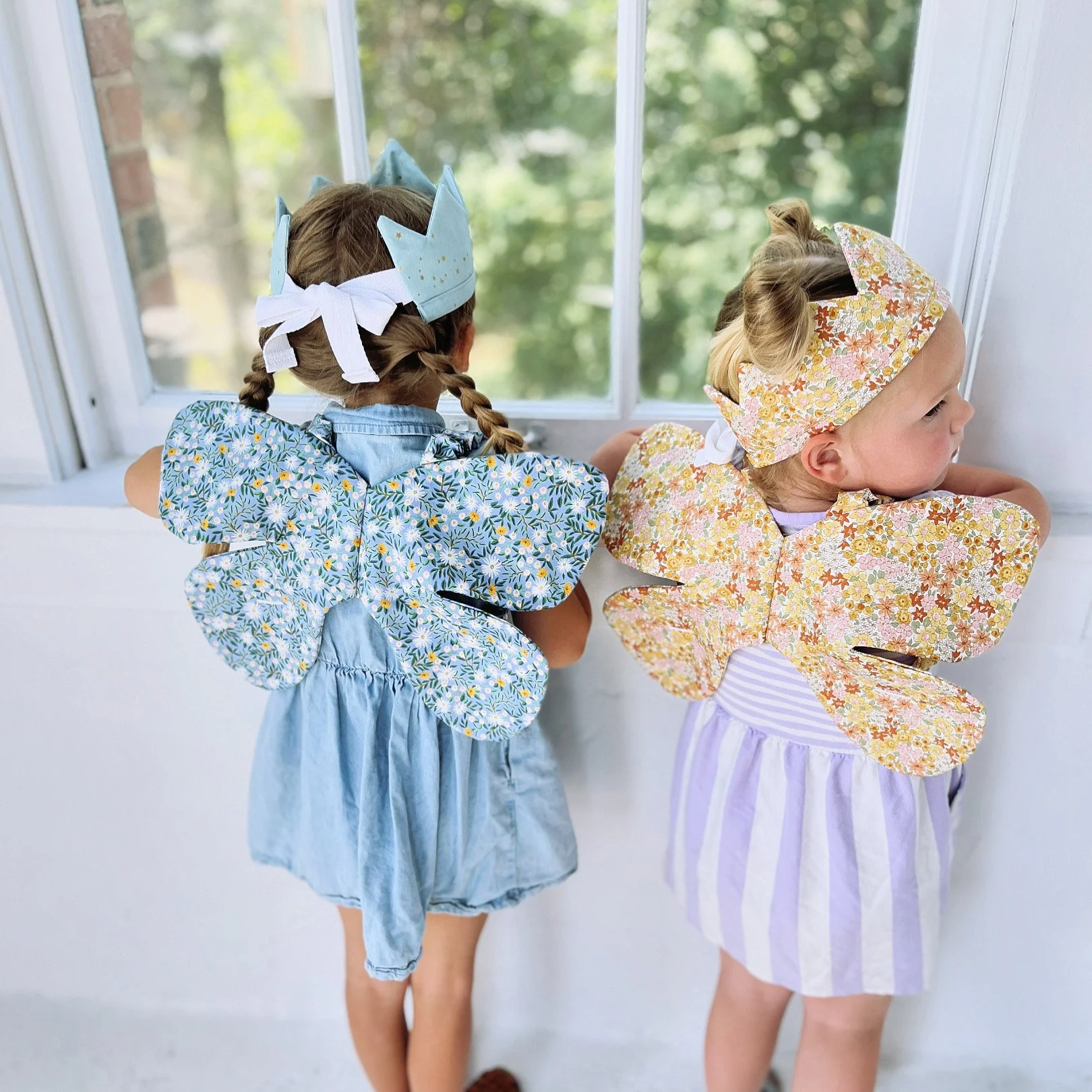 Two young girls dressed in floral outfits with butterfly wings and headbands, looking out of a window.