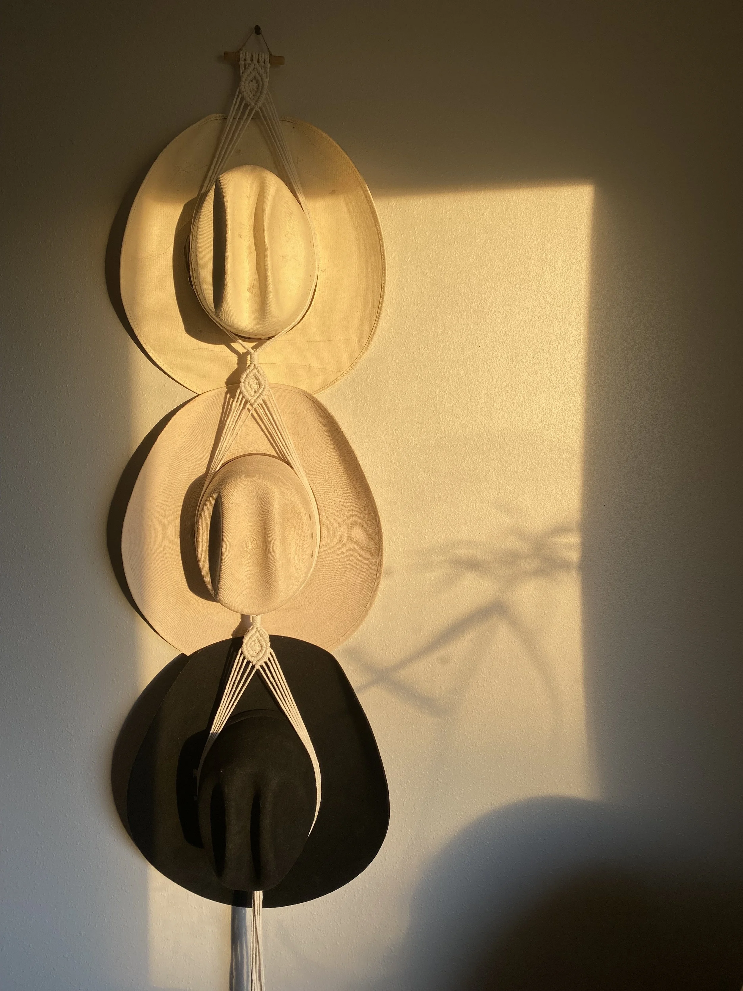 Three hats hanging on a wall with an artistic macrame holder. The top hat is a tan cowboy hat, the middle one a beige fedora, and the bottom a black wide-brimmed hat. Sunlight casts a shadow on the wall.