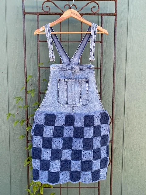 Denim bib skirt with a checkered crocheted bottom hanging on a wooden hanger against a green fence with some climbing plants.