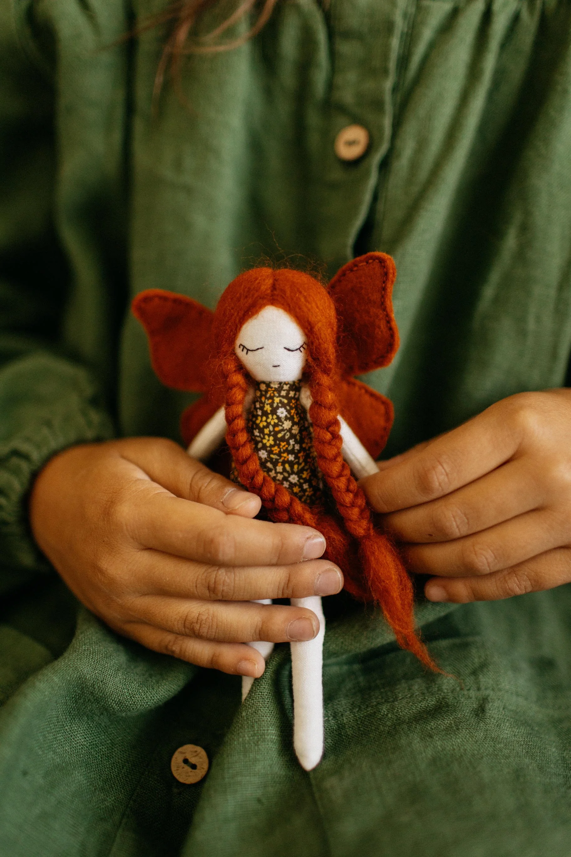 Person in green shirt holding a small doll with orange braided hair, butterfly wings, and a floral dress.