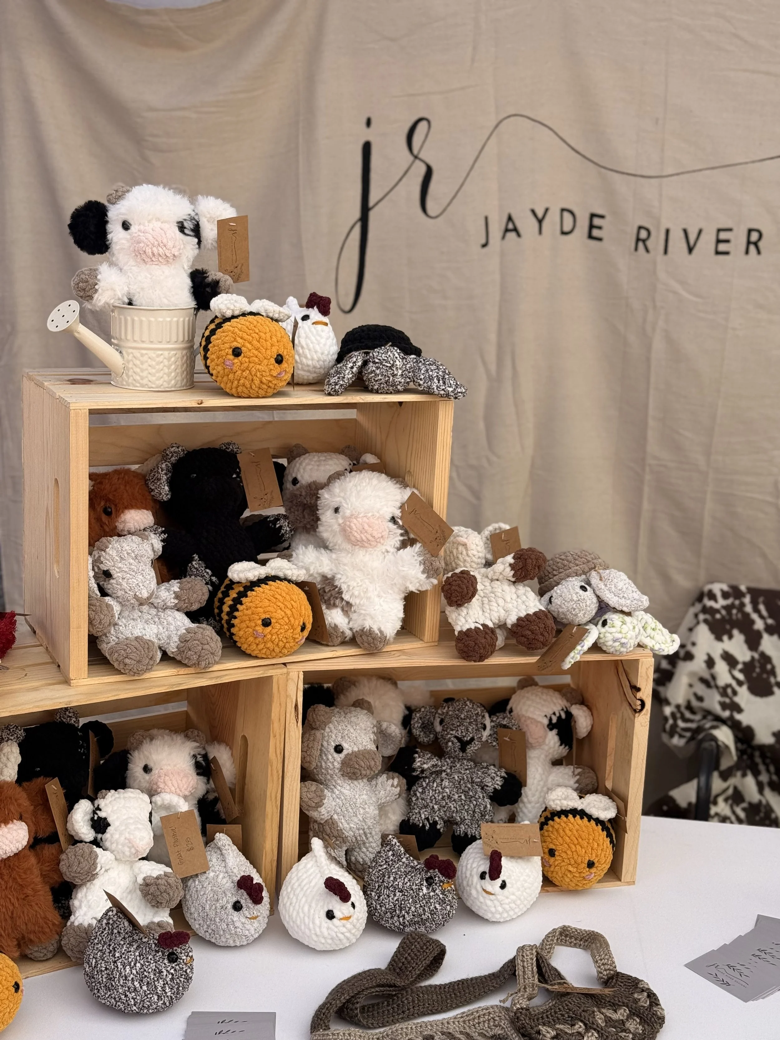 Display of various crocheted stuffed animals, including sheep, chickens, and a dog, arranged on wooden shelves and a table at Jayde River craft fair.