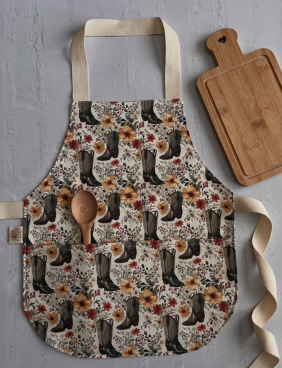 A floral apron with cowboy boots and flower pattern, a wooden spoon in a front pocket, on a gray surface alongside a wooden cutting board.