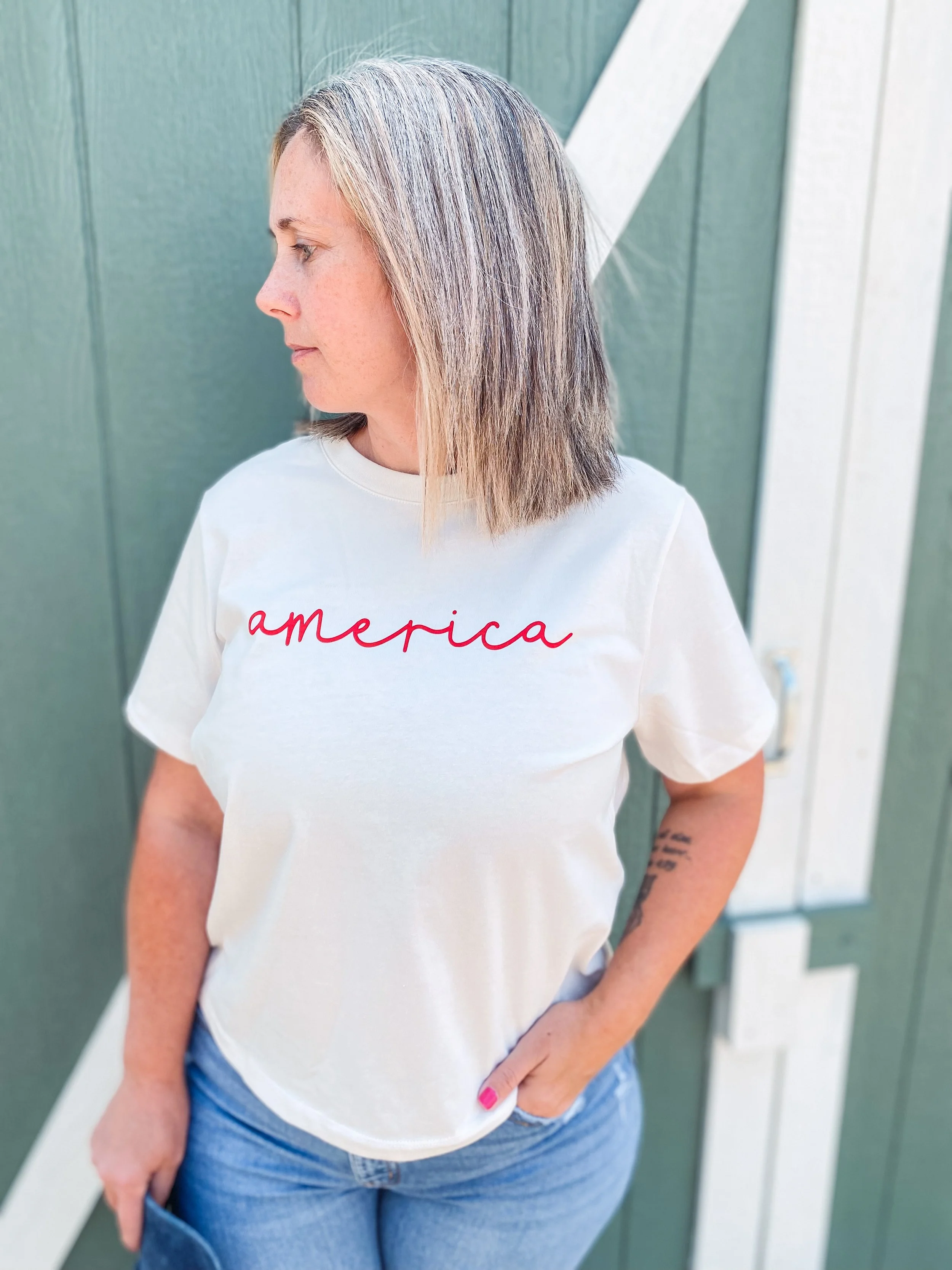 A woman with shoulder-length straight hair wearing a white t-shirt that says 'america' in red cursive and blue jeans, standing with her left hand in her pocket in front of a green wooden wall.