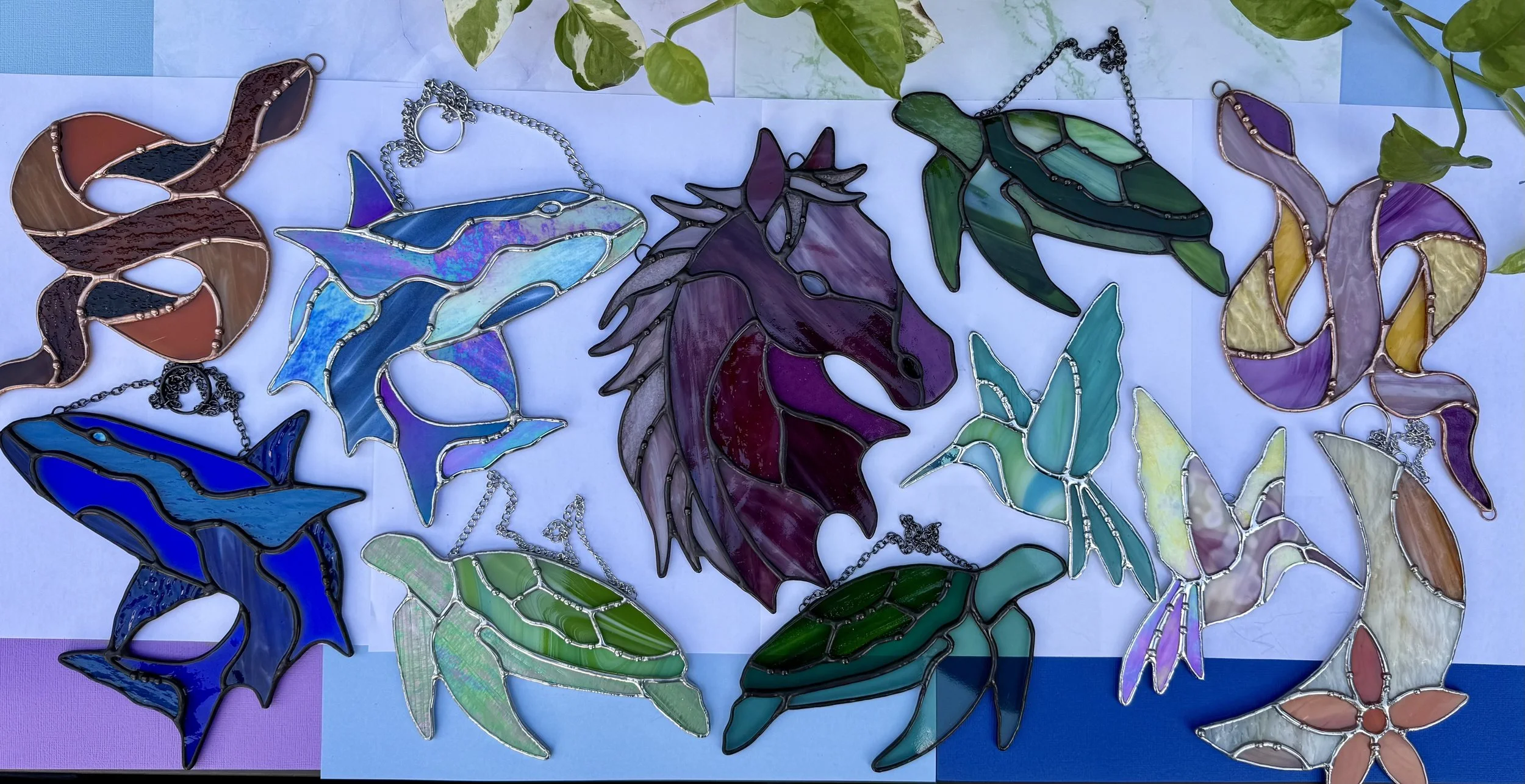 Colorful stained glass art pieces shaped like sea creatures including dolphins, a whale, a seahorse, a turtle, and a butterfly, displayed on a multicolored background. Green leaves are visible at the top.