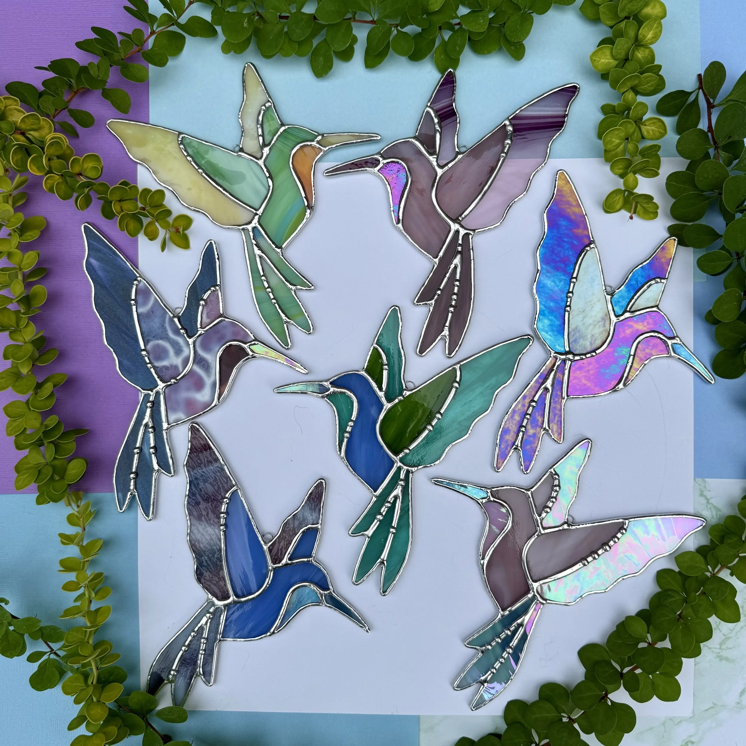 Colorful stained glass hummingbird artwork surrounded by green leaves on a backdrop of pastel-colored paper.
