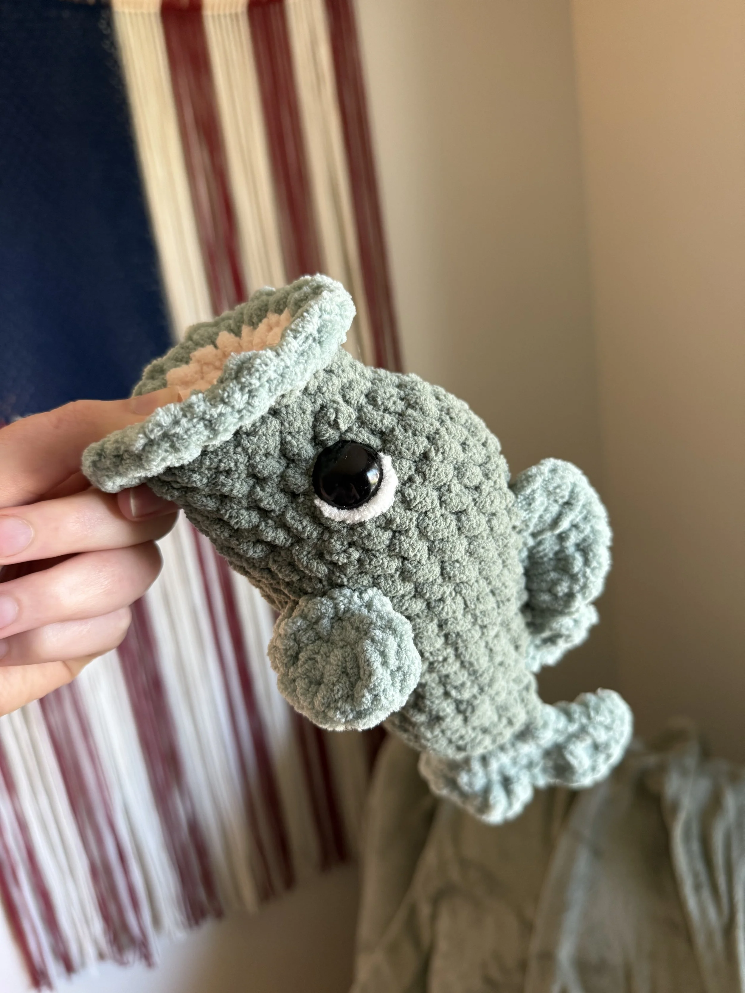 Close-up of a gray plush toy gecko with big black eyes, held by a person's hand, in front of striped curtain and beige wall.