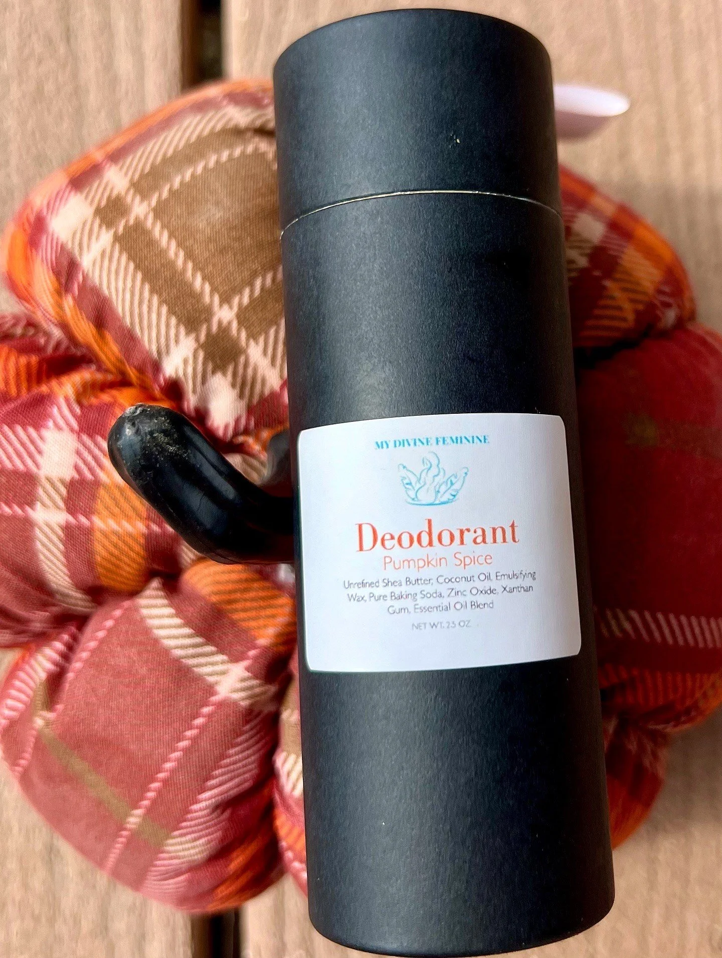 A black cylindrical container with a white label reading 'Deodorant Pumpkin Spice' and ingredients listed below, held in front of a pink, orange, and red plaid wrapped gift.