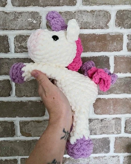 A hand holding a crocheted unicorn plush toy with a brick wall background.